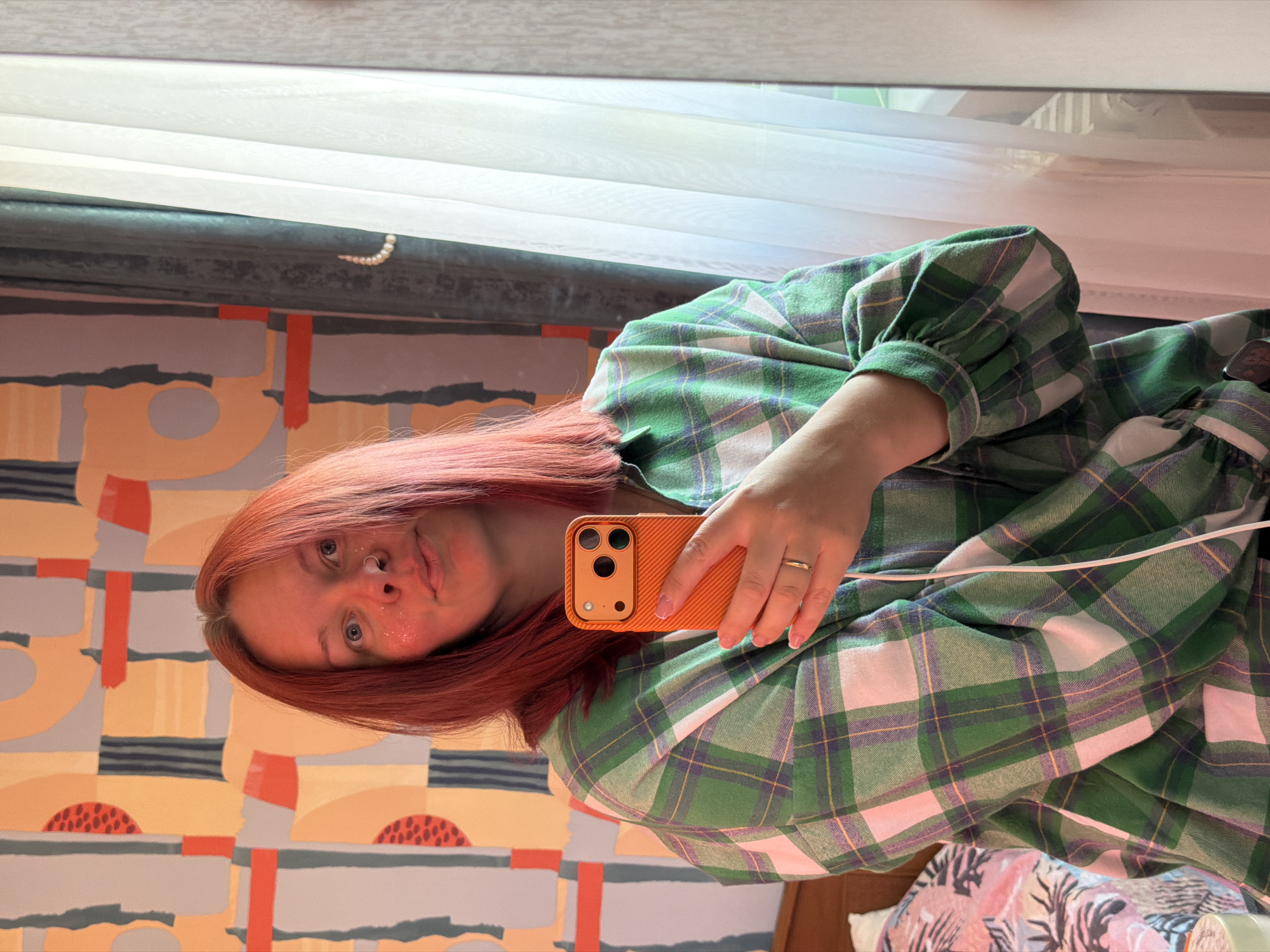 A woman with reddish-brown hair is taking a selfie in a mirror. She's wearing a green and white plaid shirt or dress. She's holding an orange smartphone. A window with white curtains is visible behind her. The wall behind her has a colorful geometric pattern. The location is indicated as Chaikovsky, Russia.