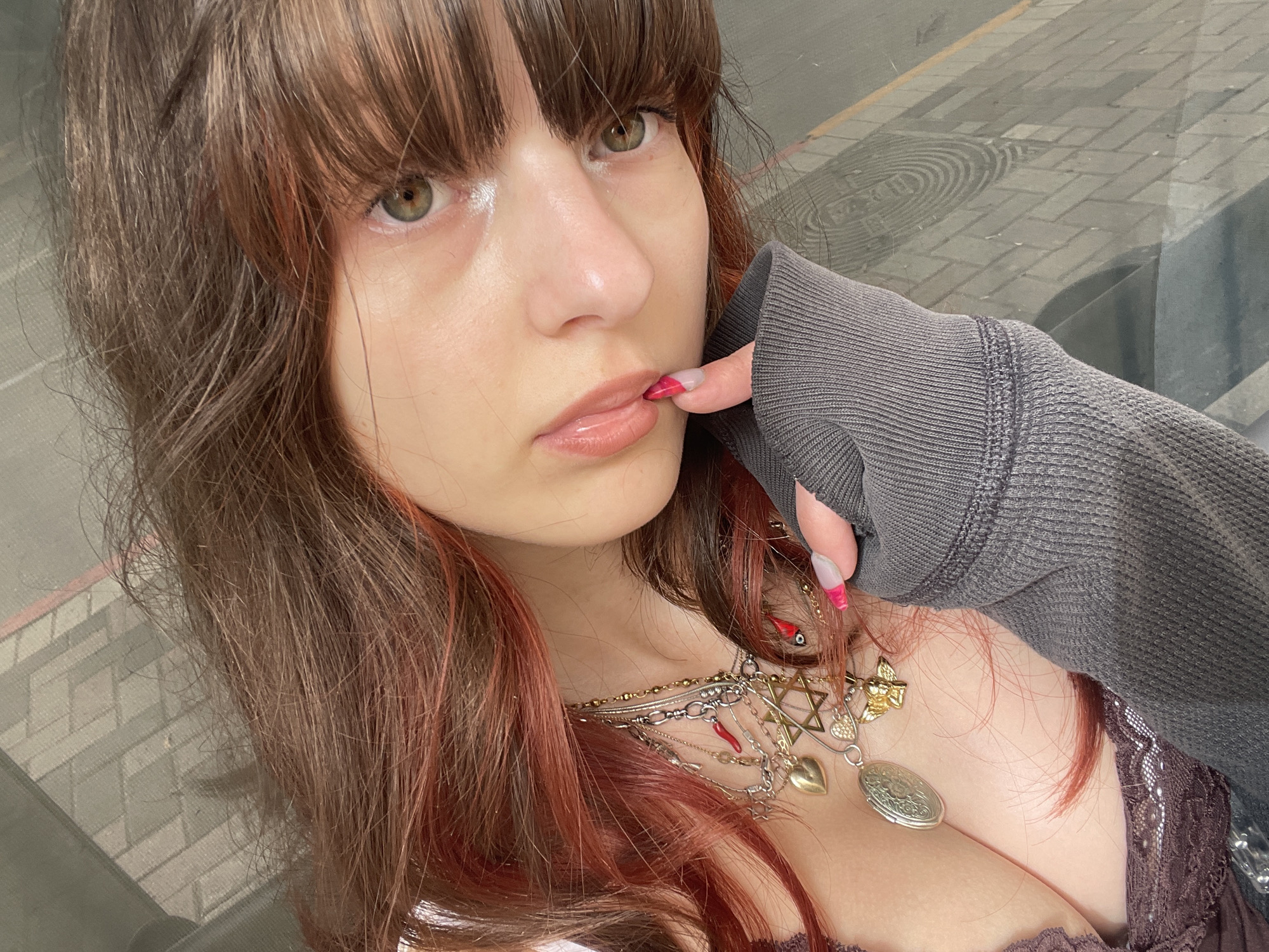 A young woman with brown hair and bangs is posing indoors near a window. The woman's hair has reddish-brown highlights. She is wearing a gray sweatshirt and a brown top with lace trim. She is wearing several necklaces. Her fingernails are painted with a red-tipped design. The background appears to be a cityscape through a window. The image does not provide enough information to confirm the location as Petah Tikva, Israel.