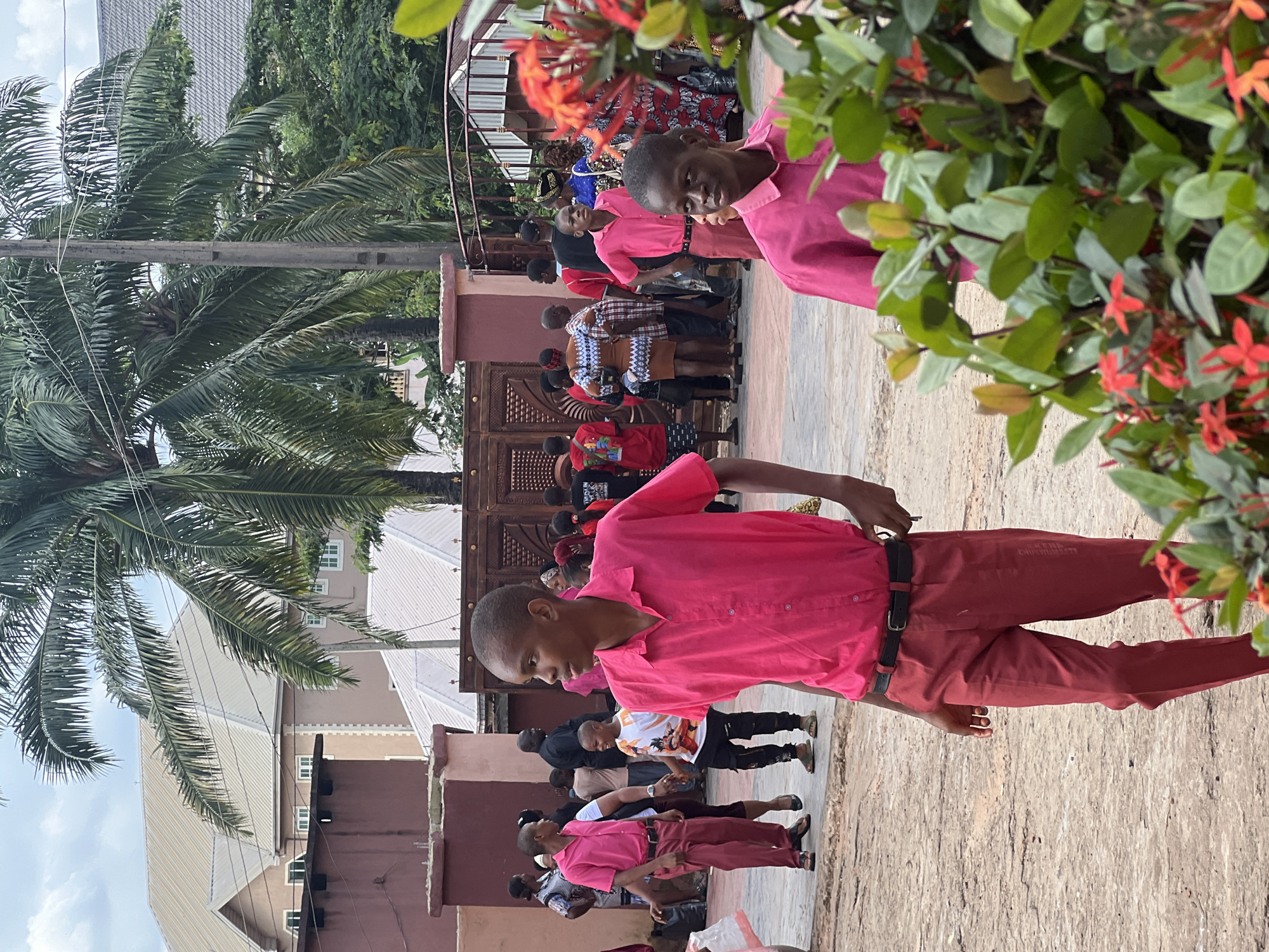 The image captures an outdoor scene on a cloudy day in Nnewi, Nigeria. In the foreground, a young boy in a bright pink collared shirt and matching trousers stands facing right, looking away from the camera. Behind him, another boy in a similar pink shirt and trousers is walking away from the camera. Several other individuals, including adults and children, are visible in the midground, some wearing pink shirts and others in diverse attire. They are gathered near a large, ornate wooden gate and a concrete wall. In the background, palm trees and buildings with light-colored roofs suggest an urban environment. The ground appears to be a dirt or gravel surface. A flowering bush with red blossoms is in the extreme foreground, partially obscuring the view.