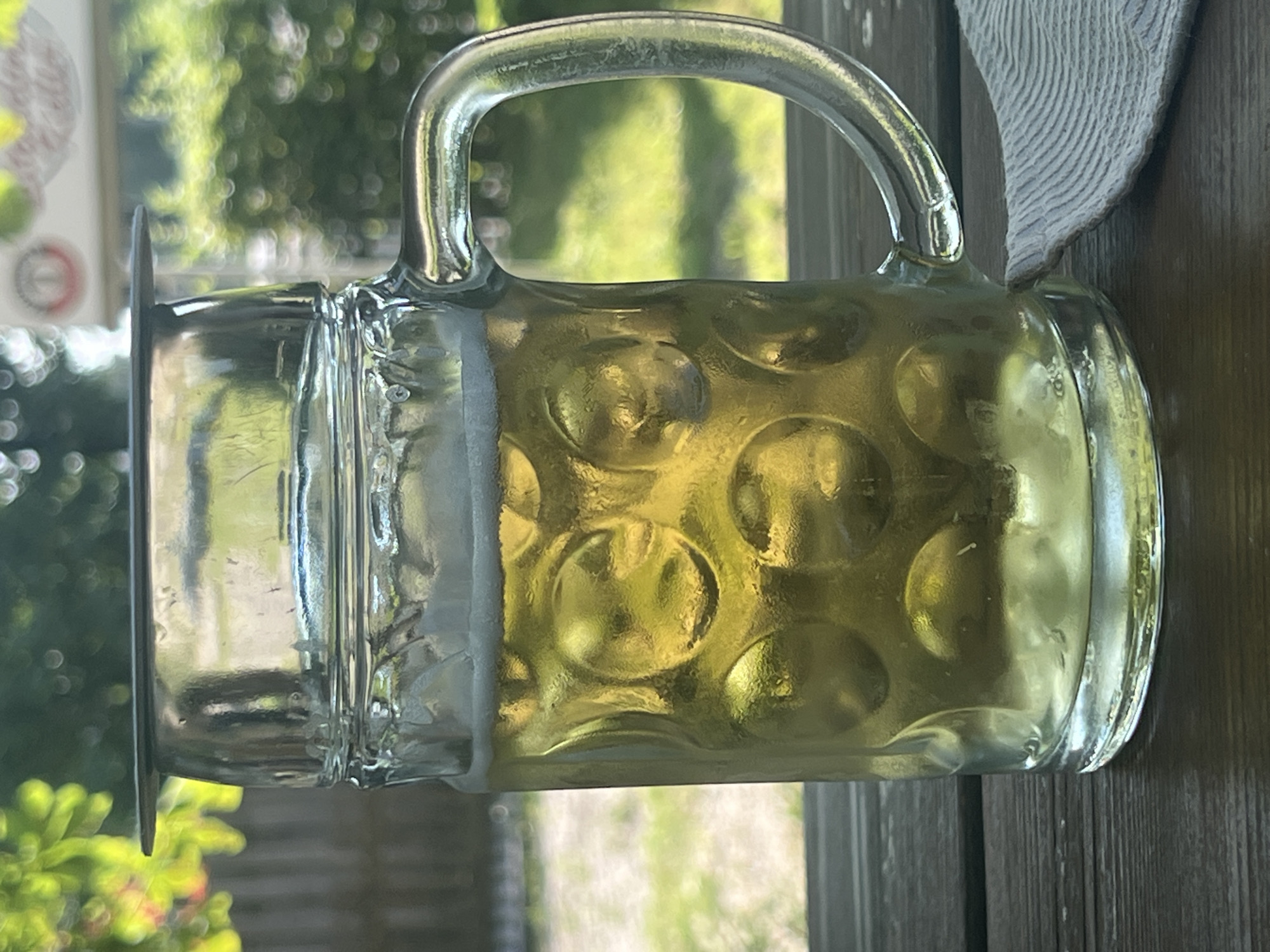 A close-up, eye-level shot features a glass beer mug filled with golden lager, topped with a frothy white head. The mug has a thick, ridged design on the lower half and a clear handle. It sits on a dark wooden table, with a grey and white striped napkin to the right. The background is blurred, showing greenery and a wooden fence, suggesting an outdoor setting like a beer garden or Biergarten, possibly in Moosburg, Germany, given the context. The overall lighting indicates daylight.