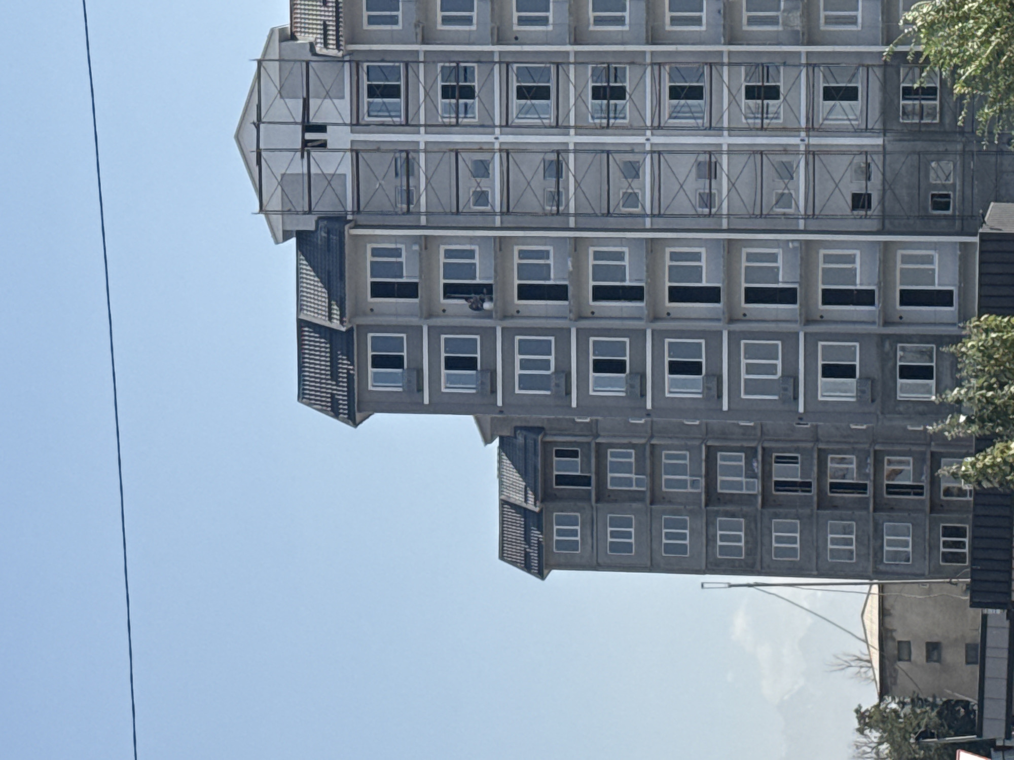 A wide shot of a partially constructed building in Bishkek, Kyrgyzstan, against a clear blue sky. The building is multi-story, with a gray facade and dark-tiled roofs. Scaffolding is visible on the right side of the building, indicating ongoing construction. Several windows are present on each floor, with some appearing to be open. In the distance, faint mountain ranges can be seen. To the left of the main building, a smaller, older structure with a beige facade is visible. Trees and foliage are present in the foreground and along the bottom of the frame. The overall impression is a scene of urban development.