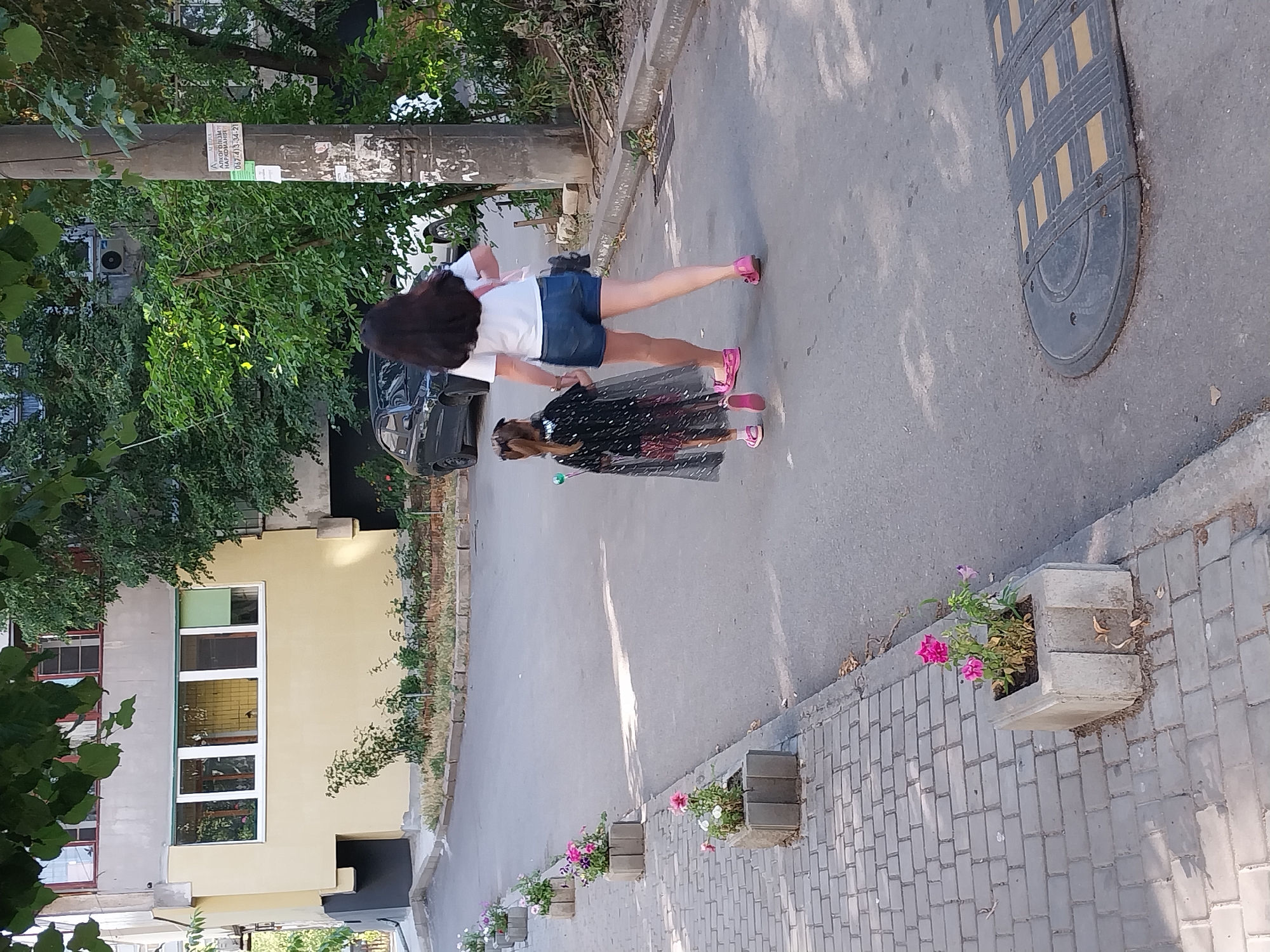 This image depicts a street scene in Dnipro, Ukraine, on a sunny day. Two individuals, a woman and a young girl, are walking away from the camera on a paved street. The woman is wearing a white t-shirt and denim shorts, with her dark hair long and flowing. She is carrying a pink bag on her shoulder. The girl, in front of the woman, is wearing a black cape with silver stars, a black and pink skirt, and pink sandals. She is holding a sparkling wand.
The street is lined with concrete planters containing flowers, with a sidewalk made of grey paving stones on the left. The background shows a yellow building with several windows, surrounded by trees and vegetation. A dark-colored car is parked on the street, and a speed bump is visible on the road in the foreground. A utility pole with posters is also present on the right side of the image. The overall impression is of a typical urban environment in Dnipro during daylight hours.