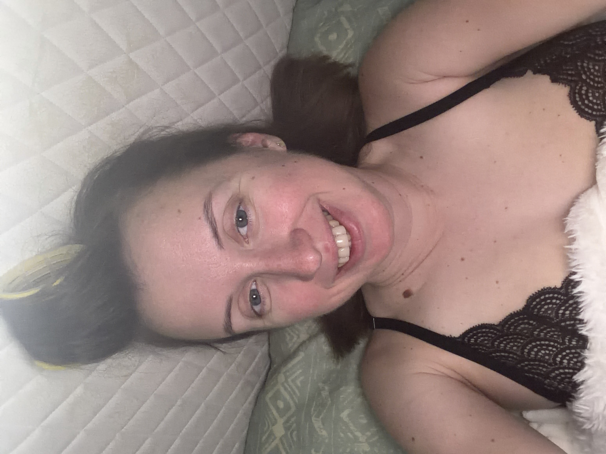 A woman with brown hair styled in rollers is lying in bed. She is wearing a black lace bra and a white furry blanket. The background shows a quilted headboard and a patterned pillow. The scene is indoors, likely a bedroom. There is no visible indication of the city of Bila Tserkva, Ukraine.
