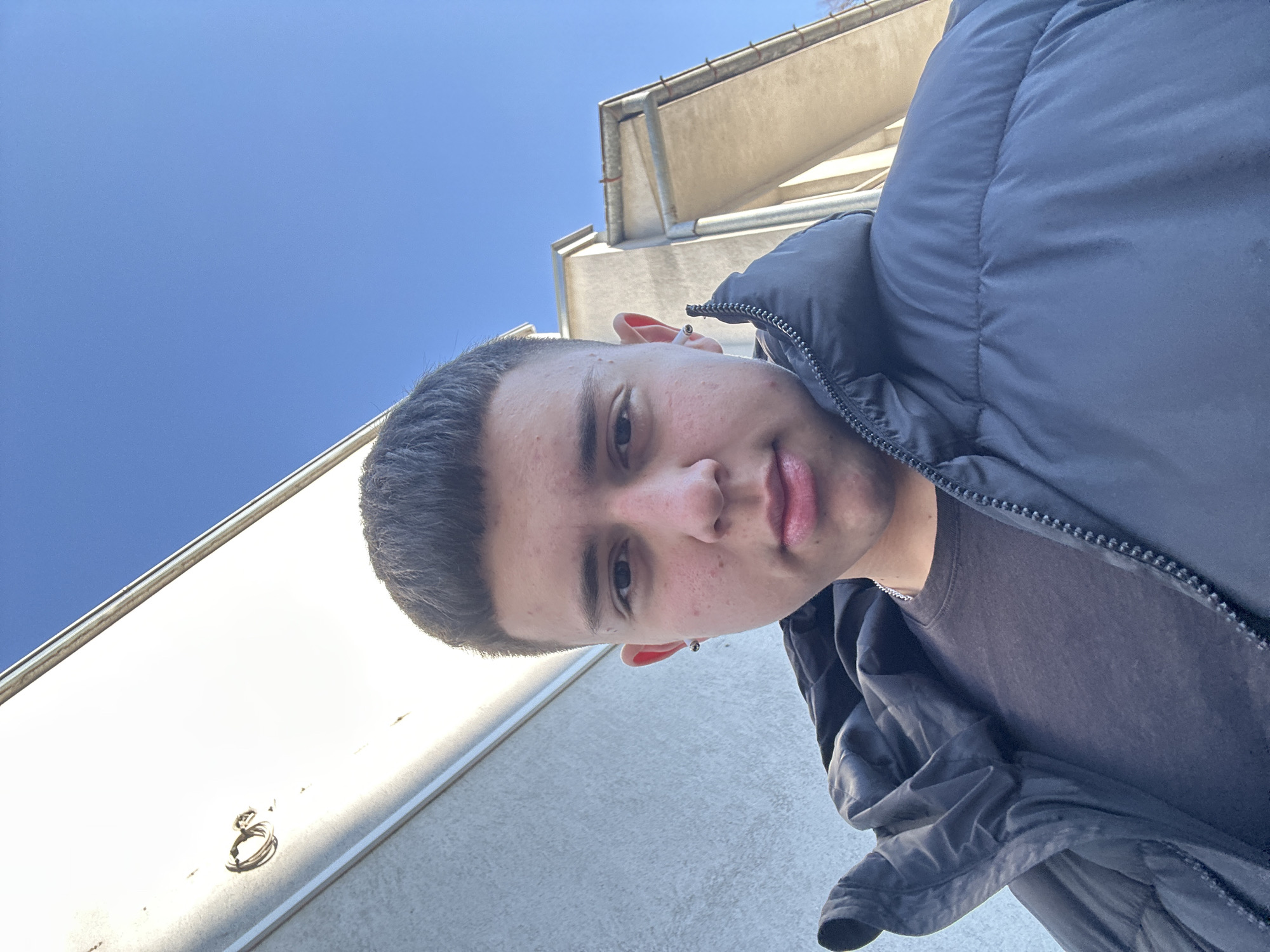 A medium shot of a young man wearing a dark puffer jacket and a dark t-shirt. The background includes a clear blue sky and parts of a building, possibly located in Zlatograd, Bulgaria. The image is taken from a slightly low angle, looking up at the man. The man has short dark hair and visible facial features. The building in the background has light-colored walls and a metallic gutter.