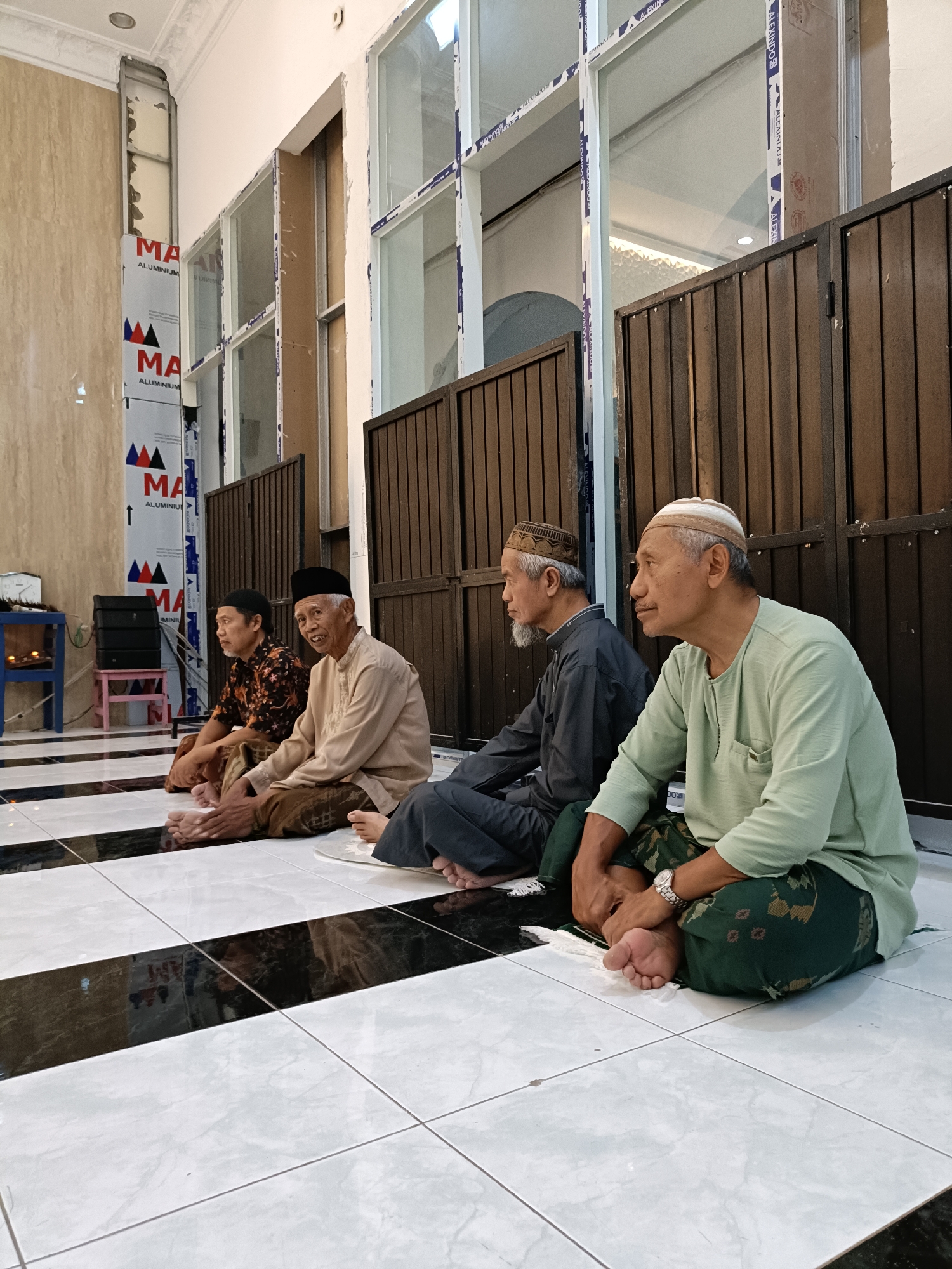 In the city of Kediri, Indonesia, four men are seated on a black and white tiled floor, facing towards the left of the frame. The man in the foreground, on the far right, is wearing a light green long-sleeved shirt and a patterned green sarong. He has a white and brown cap on his head and is wearing a watch on his left wrist. To his left, a man in a dark grey long-sleeved shirt and a patterned brown sarong is also seated. He wears a grey and white patterned cap. Next to him, a man in a light brown long-sleeved shirt and a patterned brown sarong is seated, also wearing a black cap. The man furthest to the left in the image is wearing a patterned dark shirt and a patterned brown sarong, with a black cap on his head. Behind the men, a dark brown wooden door or partition is visible. To the left of the men, a blue table with a clock and a smaller pink stool are present. The wall behind the men has vertical panels with blue and red triangular logos. The floor has a striped pattern of black and white tiles. The environment appears to be indoors, possibly a place of worship or community gathering space.
