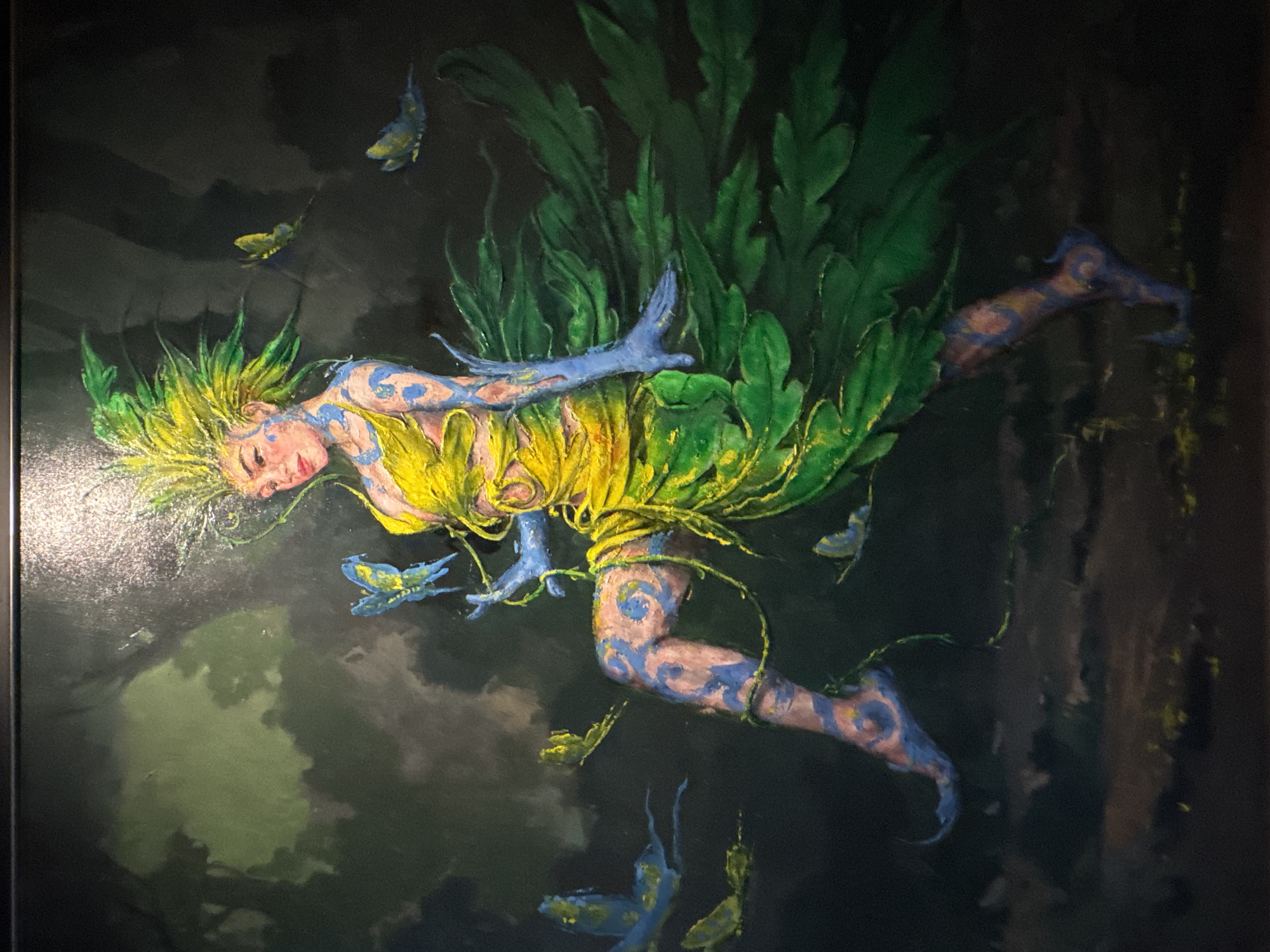 This is a vivid painting depicting a fantastical, human-like figure, possibly a fairy or nature spirit, set against a dark, lush forest background. The central subject is seen floating or flying horizontally through the air.
The figure has vibrant, spiky yellow and green hair that blends seamlessly into large, green, leaf-like formations extending from her back, resembling wings or a natural gown. Her skin is a light, rosy tone, intricately adorned with swirling blue patterns or body paint covering her arms, legs, and torso. She wears a yellow, petal-like garment around her waist. Her hands and feet are a striking blue, matching the patterns on her body. Her face is serene, with eyes closed and a soft, peaceful expression, suggesting tranquility or slumber in motion.
Numerous small creatures resembling blue and yellow butterflies or moths flutter around the figure, some appearing to emanate from or interact with the blue markings on her skin. The background is dark and shadowy, with indistinct forms of green foliage suggesting a deep, mystical forest environment. The lighting dramatically highlights the figure, making her glow against the dark backdrop.
The painting uses a rich color palette, emphasizing greens, yellows, and blues, with visible brushstrokes creating texture. The overall scene evokes a magical, dreamlike atmosphere. There is no visible text in the image. The setting's natural theme aligns with the general environment of Kuta Selatan, Indonesia.