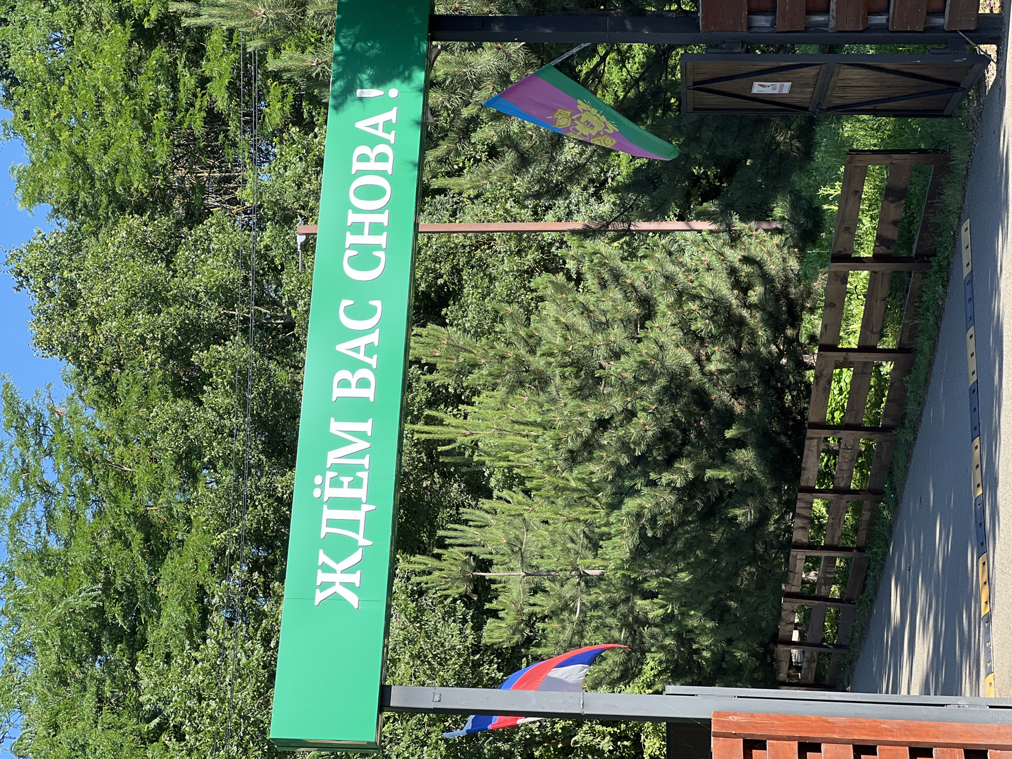 A bright green archway sign with white Cyrillic text reads "ЖДЁМ ВАС СНОВА!" which translates to "WE ARE WAITING FOR YOU AGAIN!". The sign is supported by dark gray metal posts. To the left of the sign, a Russian flag is attached to a post. To the right of the sign, a different flag with horizontal stripes of blue, pink, and green, and a crest in the center, is also attached to a post. Behind the sign, dense green trees and bushes form the background, with a clear blue sky visible above. In the foreground, a dark wooden fence runs horizontally, with a paved road in front of it. A speed bump is visible on the road. Wooden gate posts flank the entrance on either side of the fence.