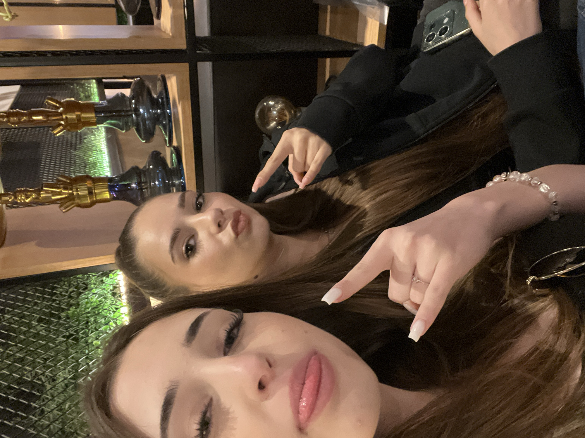 Two young women pose for a selfie indoors. The woman closest to the camera has dark hair and prominent eyelashes. The other woman, behind her, has light brown hair. Both women are pointing towards each other with their index fingers. A glass object, possibly a light fixture, is behind the women. In the background are hookah pipes on a shelf. Based on the presence of hookah pipes, the scene is likely in a hookah lounge or cafe. While it can be inferred that this scene is happening in Stara Zagora, Bulgaria, nothing in the image explicitly confirms this location.