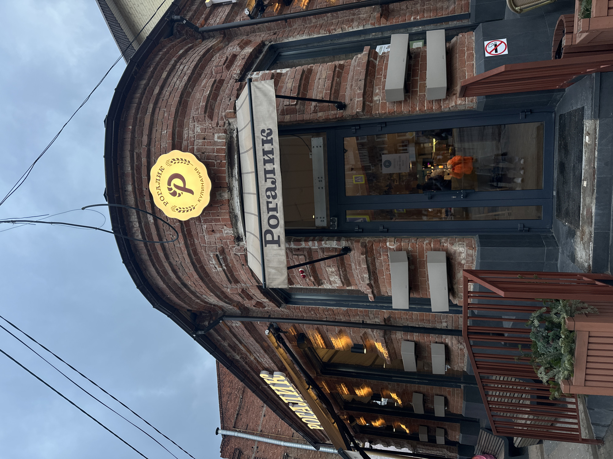 The image depicts the entrance of "Рогалик" (Rogalik) bakery in Rostov-on-Don, Russia. The building features a red brick facade with an arched doorway. A vertical beige awning displays the bakery's name in Cyrillic. A round, illuminated sign with a pretzel-like logo also bears the name "Рогалик кулинария" (Rogalik Culinary). The entrance has glass doors and a short set of stairs with a wooden railing adorned with greenery. Several white, rectangular objects are mounted on the brick wall on either side of the door. Inside, a person wearing an orange shirt can be seen. The sky is overcast. A "no smoking" sign is visible near the entrance.
