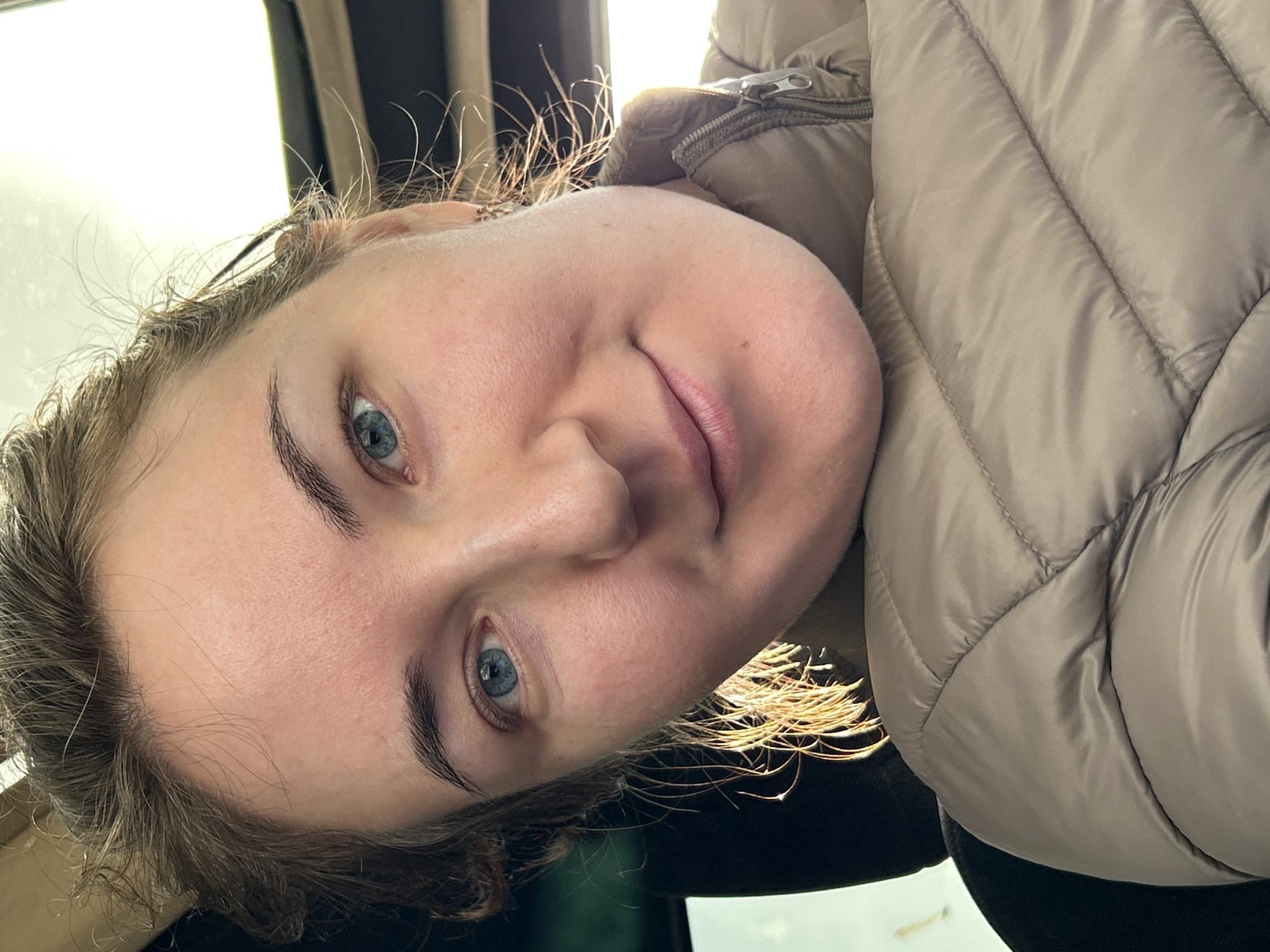 A close-up portrait features a young adult with light skin, striking blue eyes, and light brown, slightly wavy hair. The image is significantly tilted, presenting the subject's head at an oblique angle as she looks directly towards the viewer with a subtle, closed-mouth smile. Fine hairs on her face and head are visible, and her eyebrows are dark and well-defined.
She is wearing a light beige or tan quilted puffer jacket, which is visible on the right side of the frame, showcasing its textured, puffy material and a prominent zipper near the collar. A bright light source, likely from a window or the sky, illuminates the left side of her face and creates a noticeable halo effect on the individual strands of her hair. The background is largely out of focus, consisting of bright white and darker, indistinct shapes, suggesting an indoor setting or the interior of a vehicle. The lighting indicates it is daytime. No text is visible in the image. The scene is set in Chaikovsky, Russia.