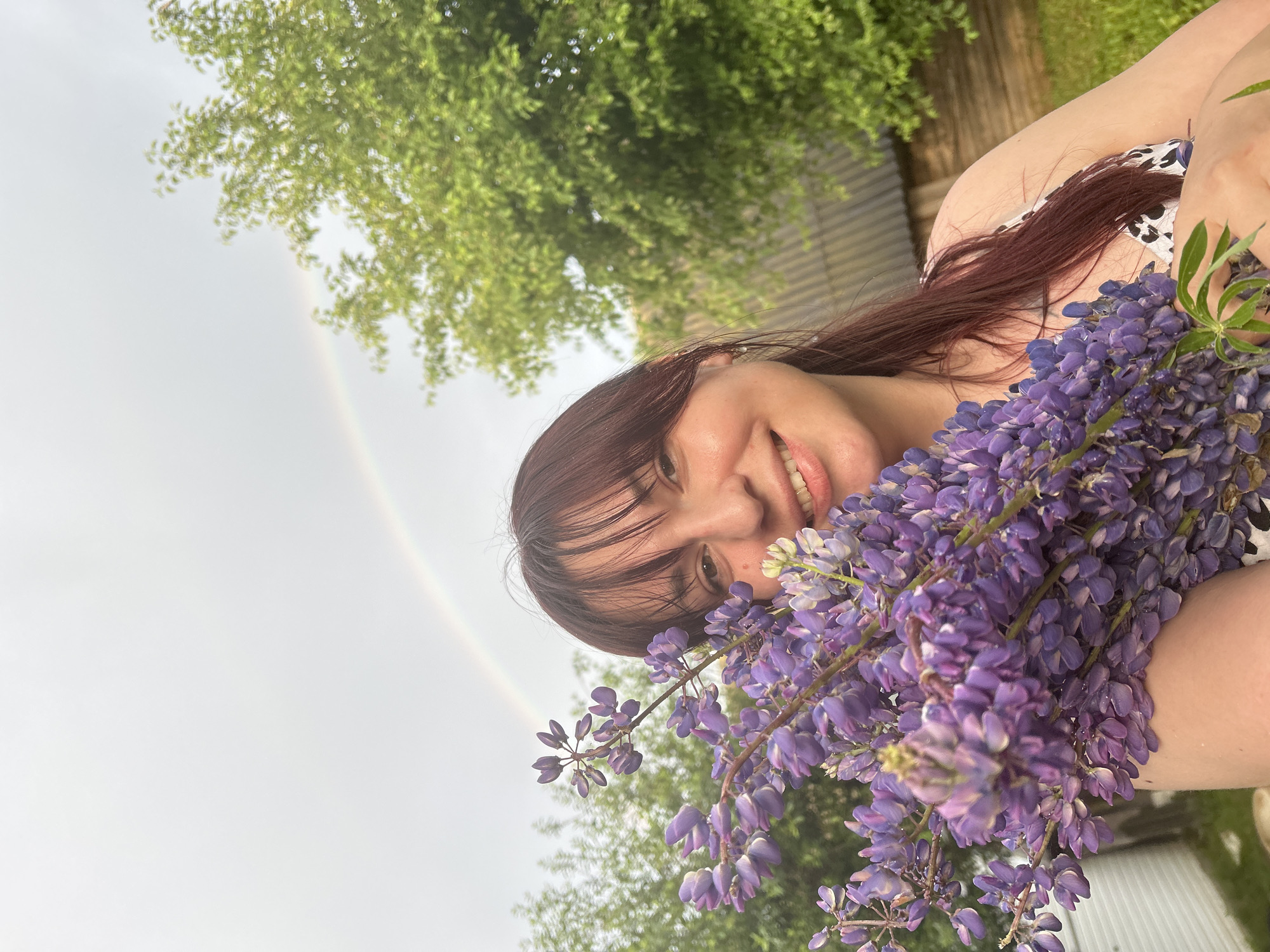 In an outdoor setting, likely in the city of Fokino, Russia, a woman with dark hair and bangs smiles at the camera while holding a large bouquet of purple lupine flowers. A faint rainbow is visible in the overcast sky behind her. Lush green trees frame the background, with glimpses of a wooden structure and fencing suggesting a rural or suburban environment. The woman is wearing a patterned dress.