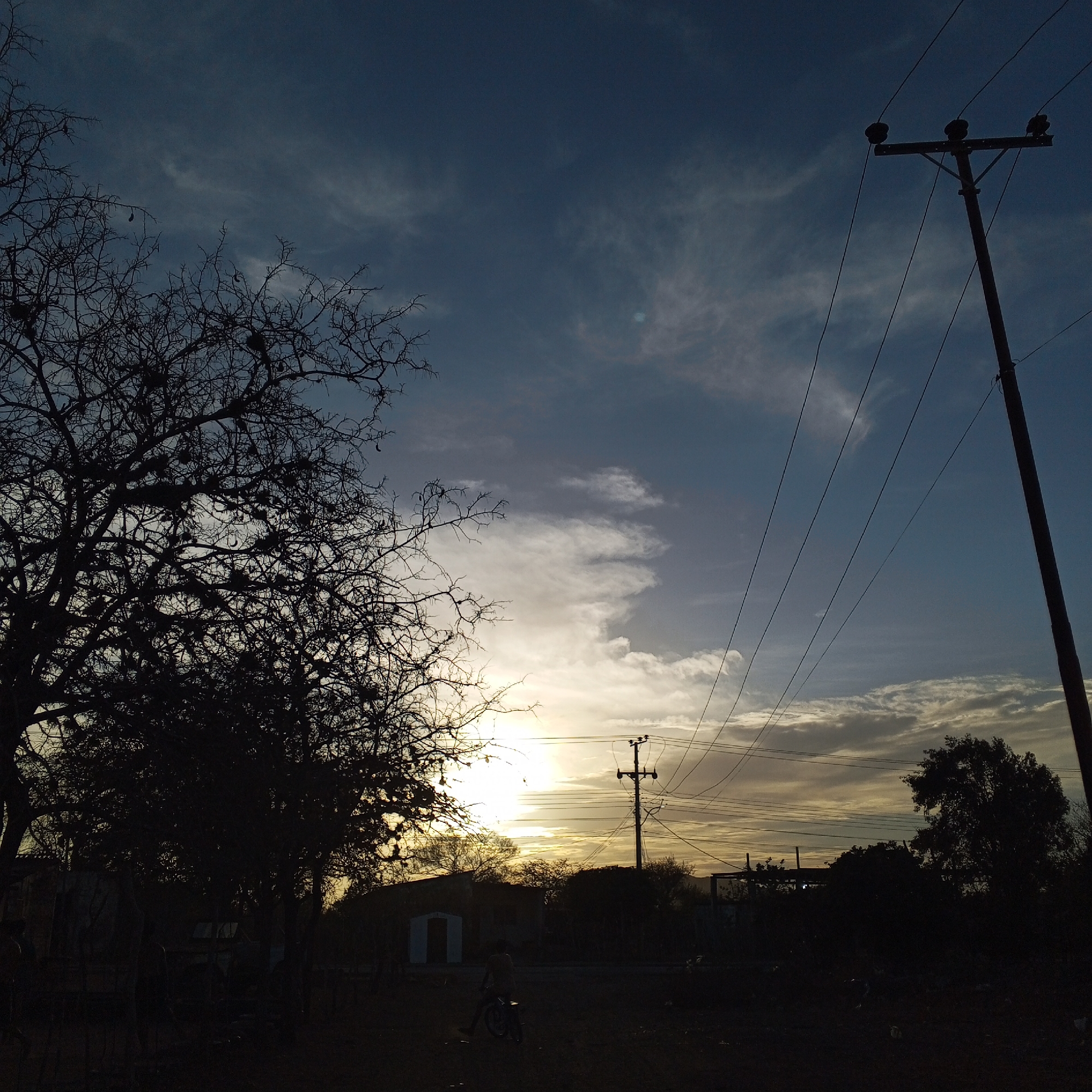 The image depicts a landscape in Unknown, Unknown, featuring a silhouetted foreground of trees and a low-lying structure, with a person on a bicycle positioned near the center. The background is dominated by a sky with varied cloud formations and the setting or rising sun creating a bright horizon. Electrical poles and wires are visible on the right side of the frame. The scene is generally dark, with the sky providing the primary source of light.