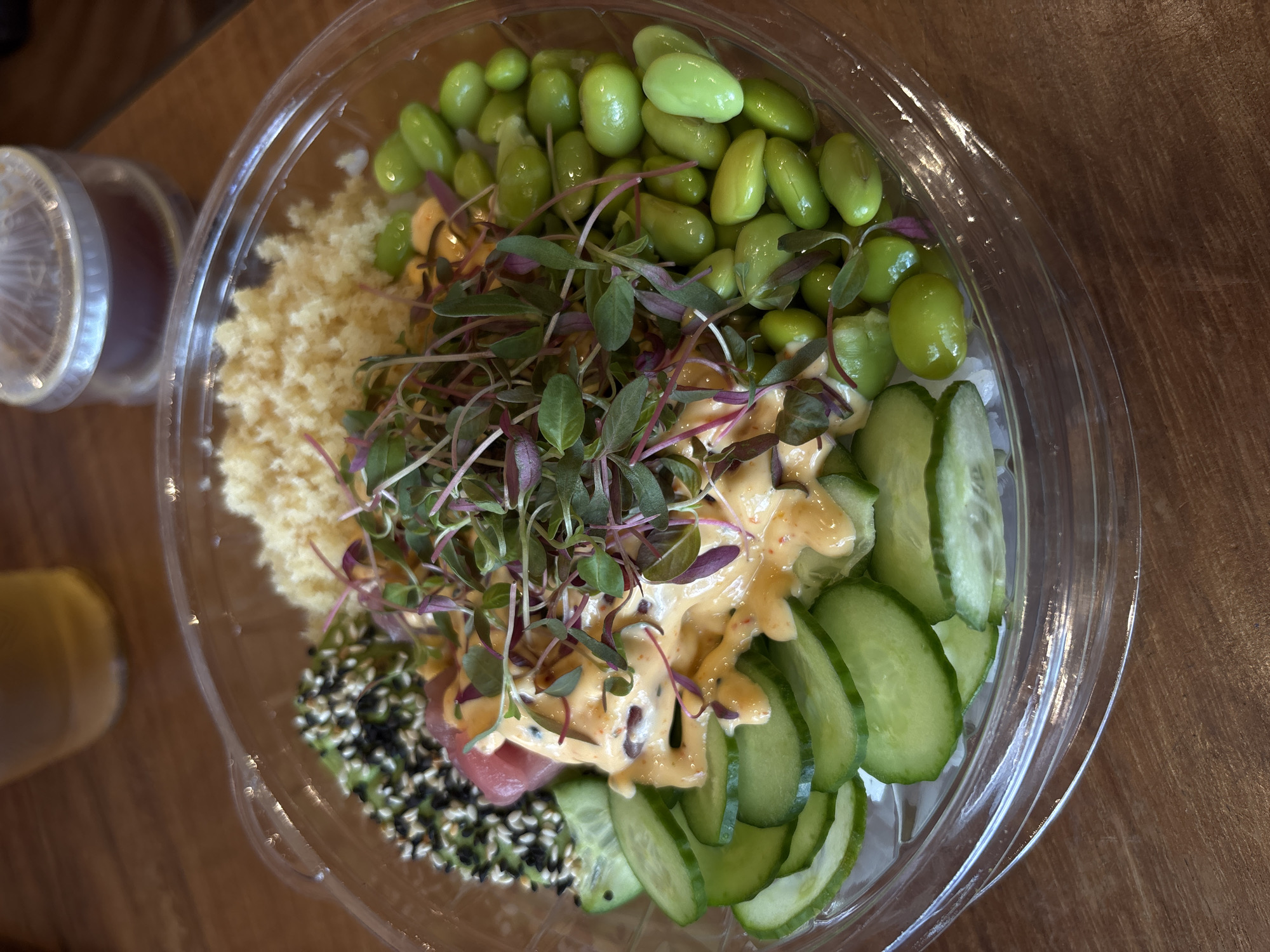 The image shows a clear plastic bowl filled with a variety of food items, placed on a wooden surface. The bowl contains edamame, sprouts, sliced cucumbers, tuna with a creamy orange sauce, black and white sesame seeds, and couscous. A small container with a lid is visible near the bowl. The location is not specified.