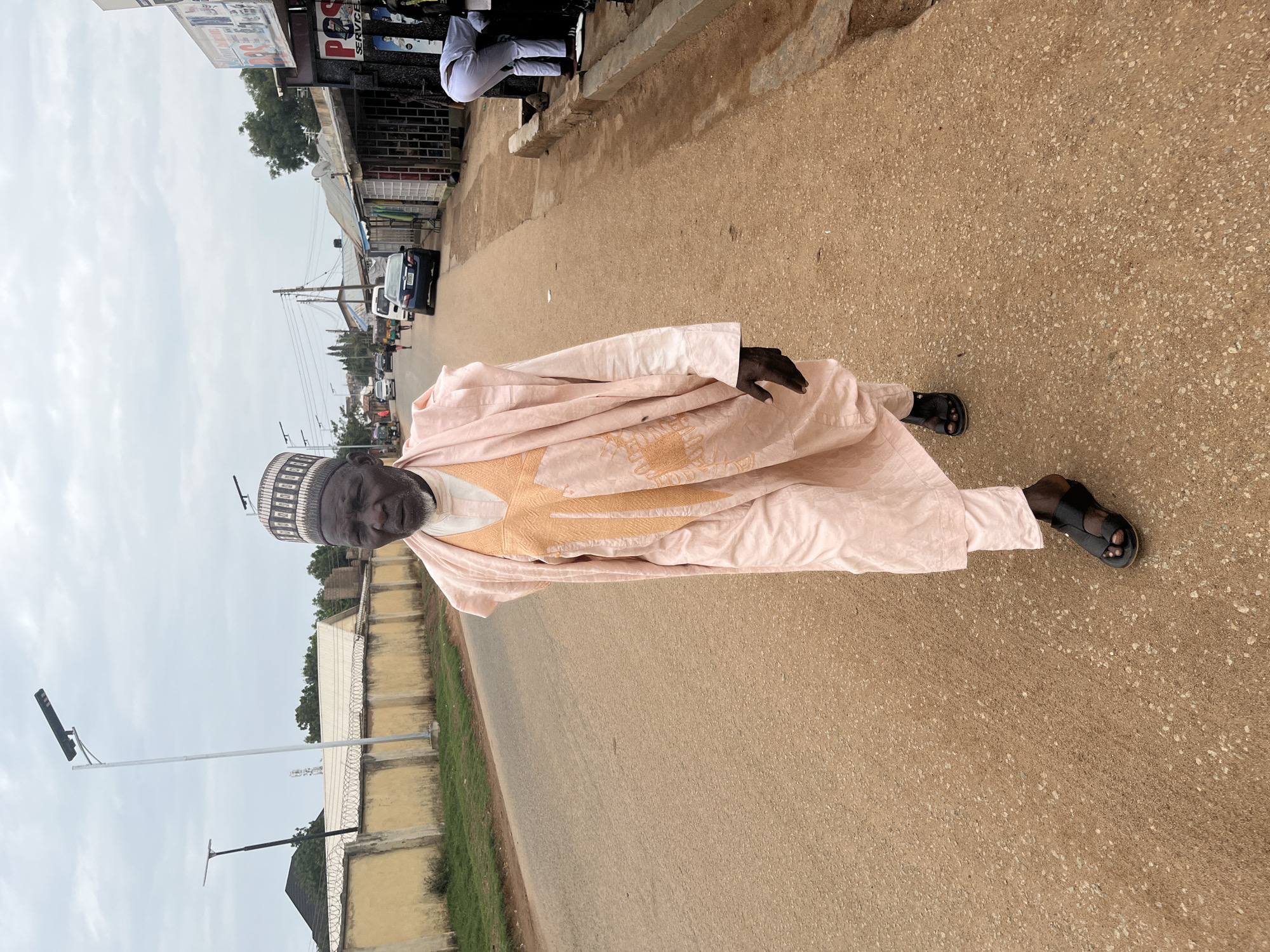 A man stands in the street in Barakin Akawo, Nigeria. He is wearing a light pink, traditional Hausa robe called a Babban Riga, and a black and white Kufi cap. He is wearing black sandals. He is standing on a paved road. In the background, there are buildings and cars, indicating an urban setting. A wall topped with barbed wire lines the edge of the road. There is a streetlight pole on the left side of the image.