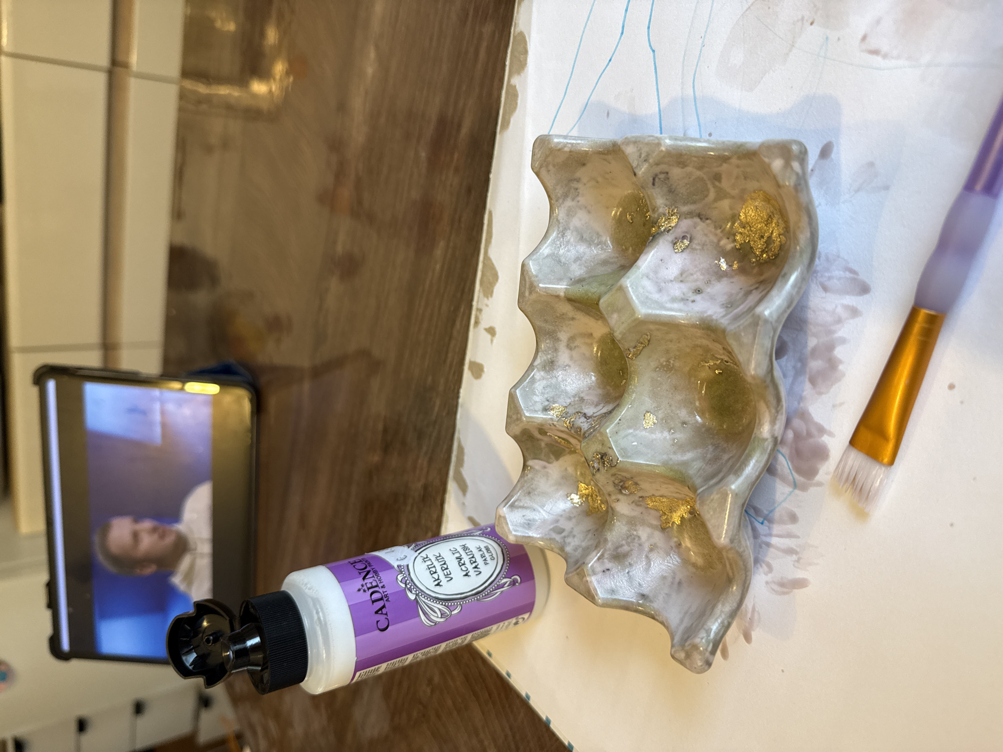 The image shows a crafting scene. On a white surface, a mold with gold leaf inclusions sits next to an open tube of Cadence acrylic varnish in a lilac-colored bottle. A paintbrush with a gold handle and purple bristles rests beside the mold. A phone stands nearby, displaying a man on screen. The environment includes a wooden surface. The location is not discernible as Zelenika, Montenegro.