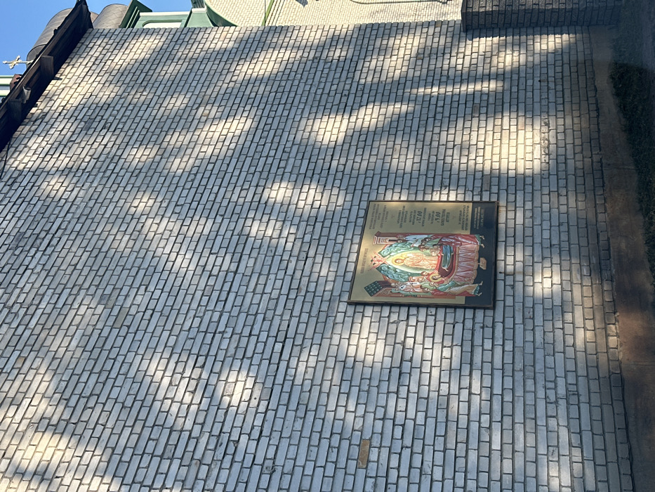 The image shows a white brick wall with shadows cast by trees and the sun. The wall is located at latitude 50.343 and longitude 30.316 in the city of Tarasivka, Ukraine. There is a framed image hanging on the wall depicting a religious scene, possibly a depiction of Jesus Christ. The frame is made of a gold-colored material. The top edge of the wall is slightly obscured by the foliage of a tree. There is no evidence of people or objects in the image.