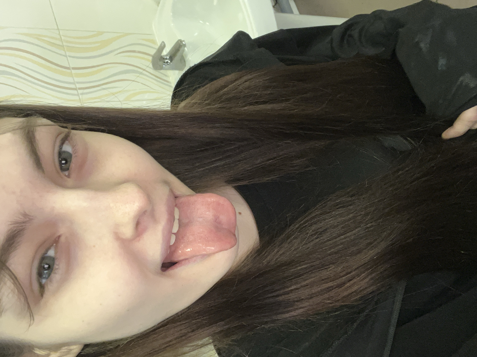 A young woman with long brown hair and pale skin is sticking out her tongue in a selfie. She is wearing a black hoodie. In the background, there is a bathroom sink and tiled wall. There is no indication in the image that this is Saint Petersburg, Russia.