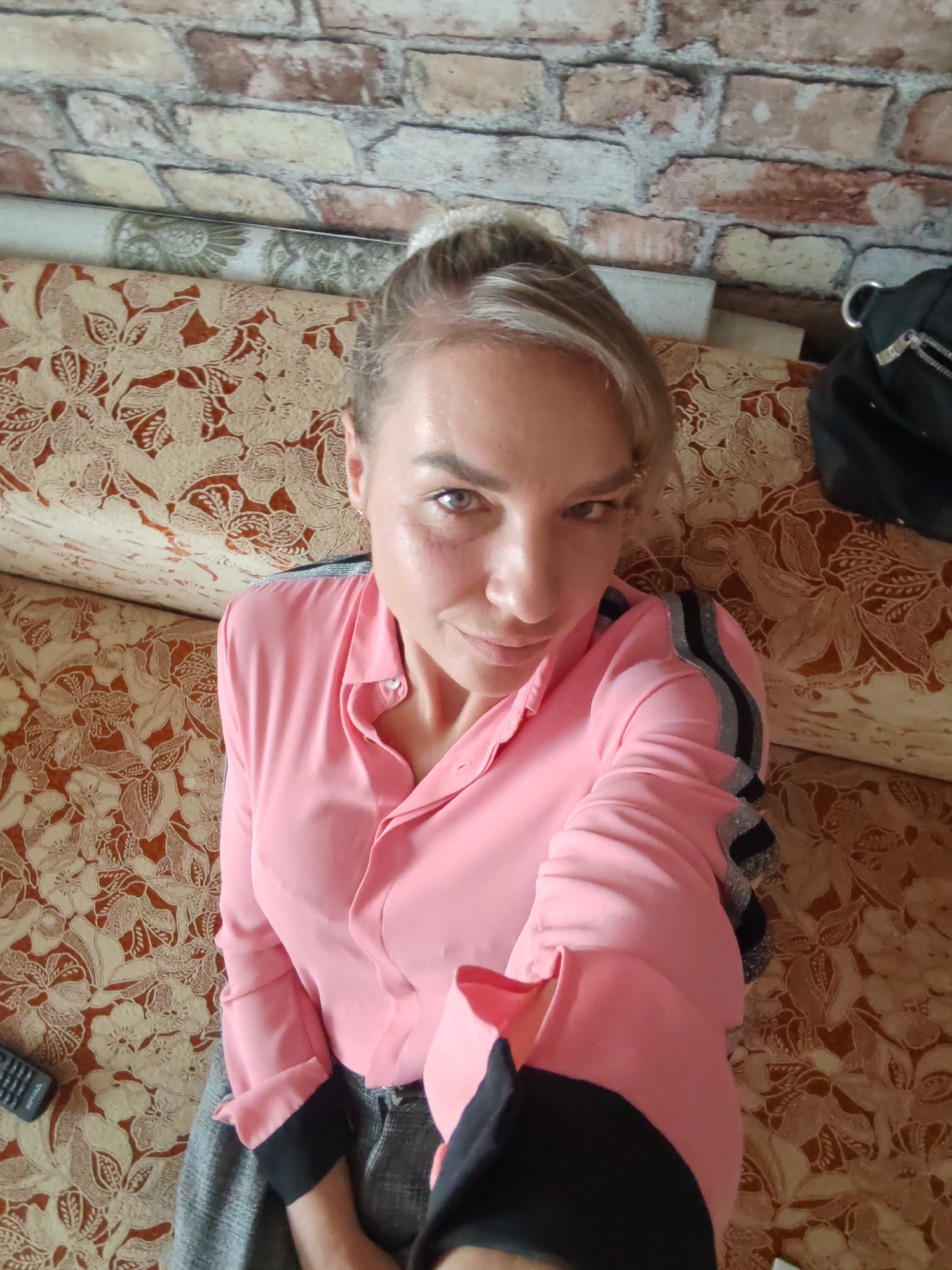 A woman with blonde hair pulled back in a bun takes a selfie while sitting on a patterned sofa. She is wearing a pink collared shirt with black and silver striped details on the shoulders and cuffs. Her lower body is out of frame, but she appears to be wearing gray pants. To the left of the sofa, a black remote control is visible. Behind the woman, a brick-patterned wallpaper can be seen. To the right, a black backpack with silver hardware rests on the sofa. The overall scene suggests an indoor setting, possibly a living room.