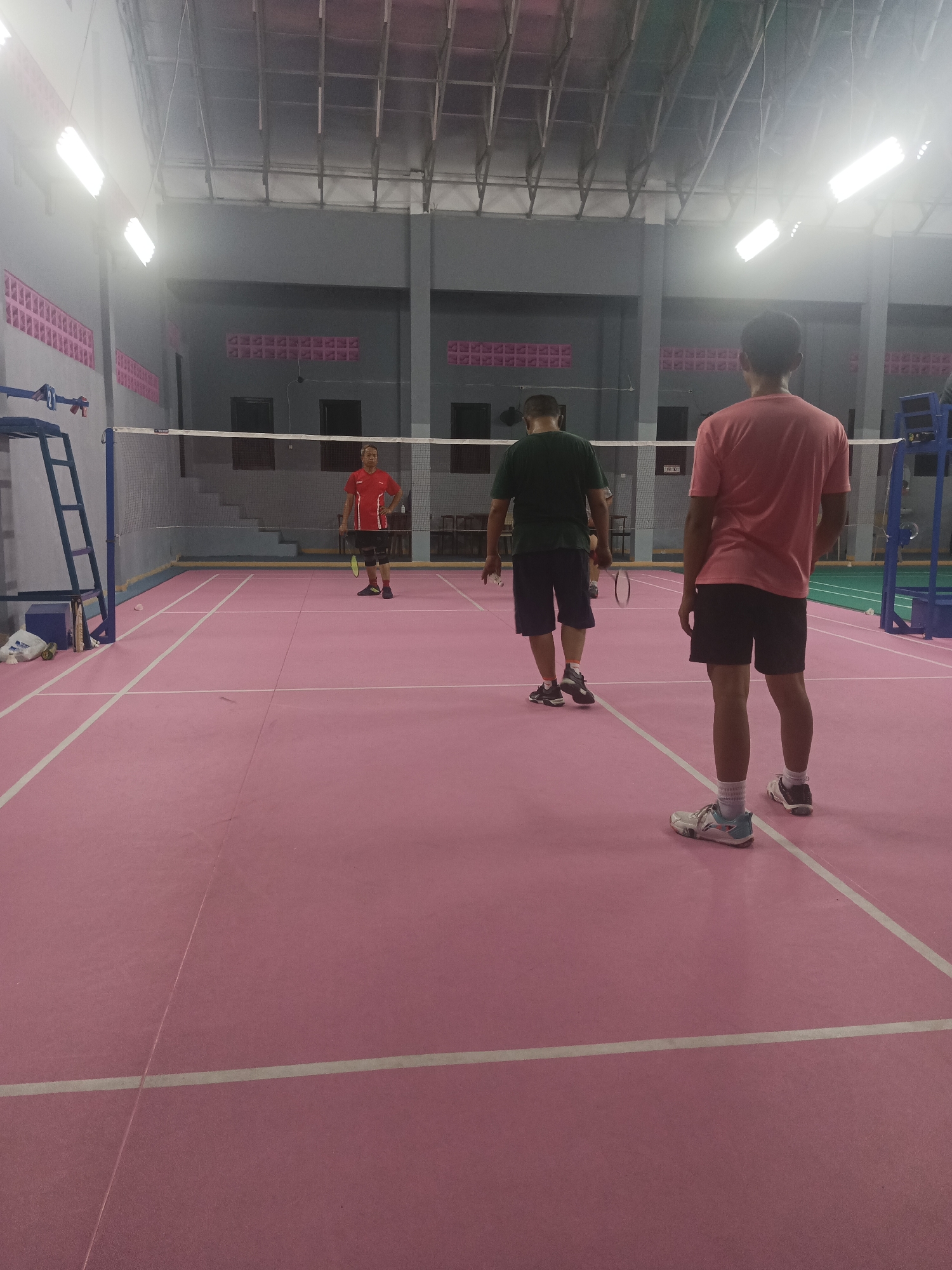 The image shows an indoor badminton court with a pink floor and white lines marking the boundaries. Three individuals are present on the court. In the foreground, on the right, a young man wearing a pink t-shirt and black shorts stands facing away from the camera, holding a badminton racket. On the left side of the court, a man in a red t-shirt and black shorts with knee pads is holding a badminton racket and appears to be in the middle of a game. Further back, in the center of the court, another individual in a dark green t-shirt and dark blue shorts is also holding a badminton racket and facing away from the camera. The court is well-lit by overhead fluorescent lights. The background shows the walls of the indoor facility, with some seating areas visible. A badminton net divides the court. The location is identified as an indoor badminton court in South Tangerang, Indonesia.