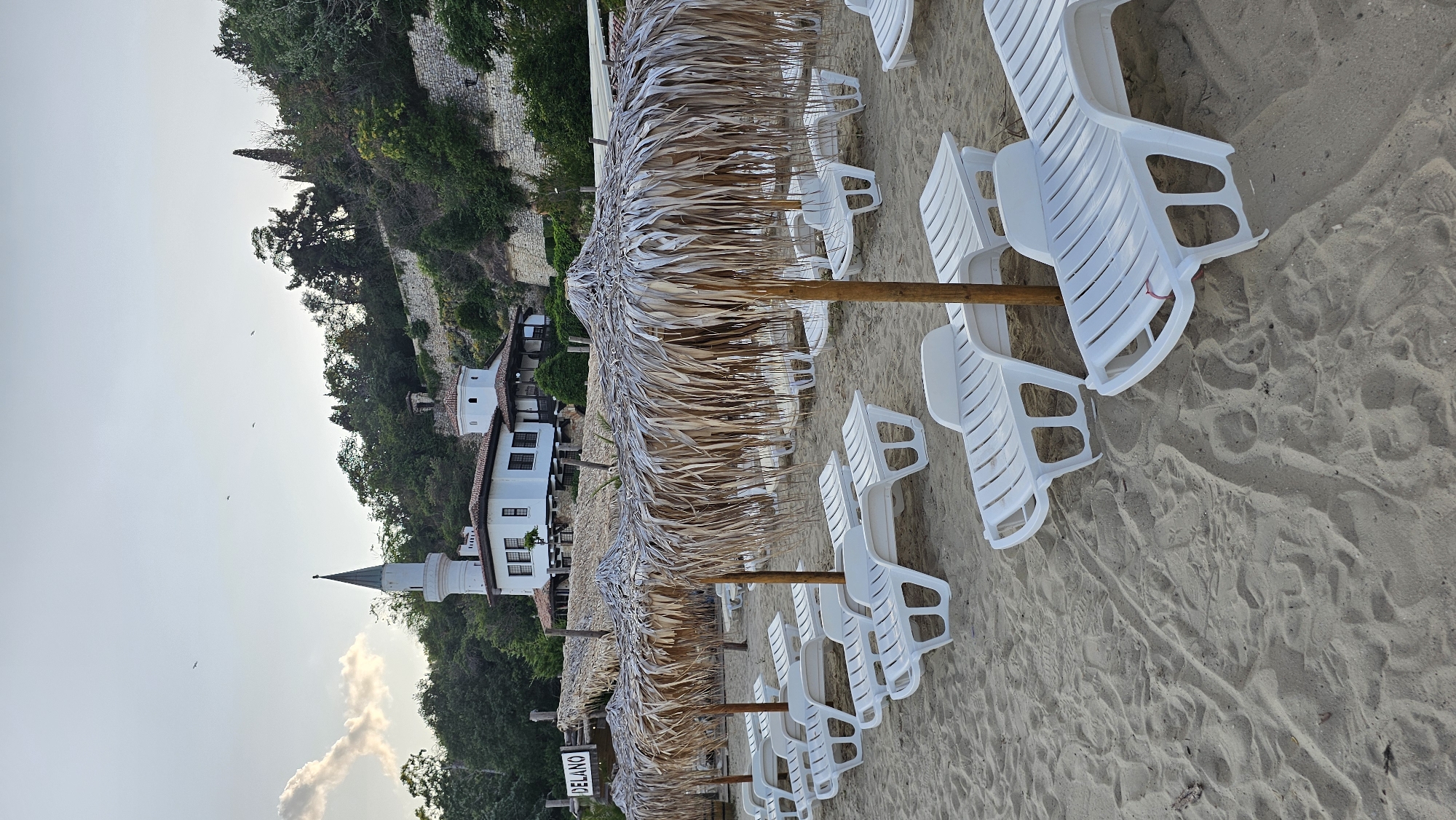 A beach scene in Balchik, Bulgaria, features a row of empty white plastic lounge chairs lined up on the sand under thatched umbrellas. In the background, a white building with red-tiled roofs and a tall, slender tower is situated on a hillside. The hillside is covered with lush green trees and stone retaining walls. The sky is overcast with a few birds visible. A sign with the word "DELANO" is partially visible on the left side of the image. The sand shows footprints and debris.