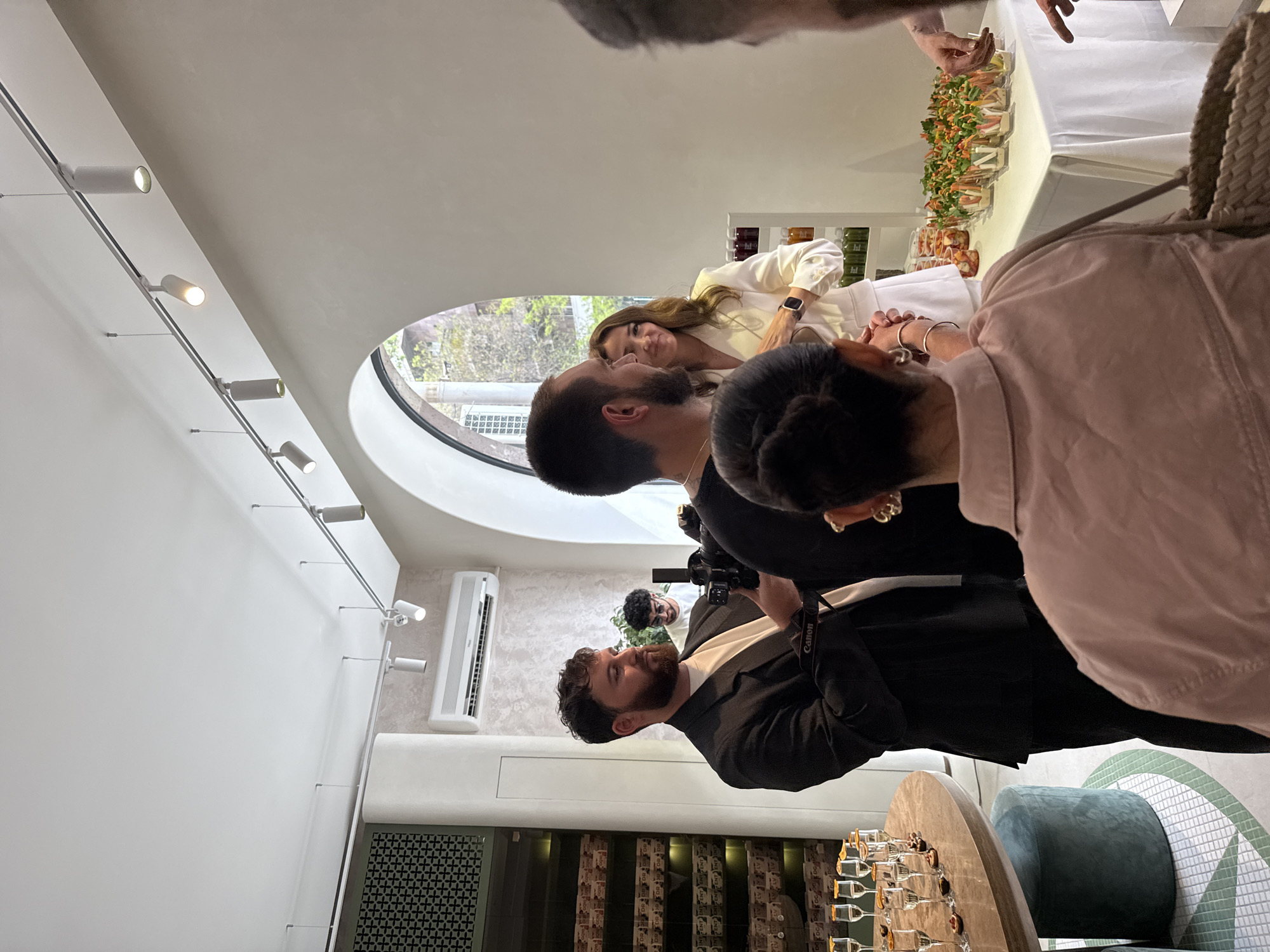 This media file captures an indoor social gathering or event in Yerevan, Armenia, set in a modern, well-lit space. The scene shows several individuals mingling and interacting.
In the foreground, from a slightly elevated perspective, a person with dark hair styled in a bun, wearing a light pink top, is seen looking towards the right side of the frame. Next to them, a bearded man in a dark suit jacket over a white shirt is holding a camera with "Canon" visible on its strap, appearing to be documenting the event. Further into the room, another bearded man is looking towards a woman with long brown hair, who is wearing a white blazer and smiling gently.
The setting features contemporary design elements, including a white ceiling with a modern track lighting system displaying several cylindrical spotlights. A distinctive large, oval-shaped window offers a view of green trees and possibly urban buildings outside, suggesting it is daytime. An air conditioning unit is mounted on a wall.
Various objects are arranged throughout the room: a round, light-colored table holds an array of empty champagne flutes and small food items, beside which sits a teal velvet ottoman. In the background, a long table is laden with numerous individual clear containers filled with colorful, fresh food items like salads or appetizers. Shelves behind this table display bottles or containers of various colorful liquids.
The atmosphere appears relaxed and pleasant, indicated by the smiling woman. The presence of a photographer suggests a formal event, such as a product launch or a celebratory gathering.