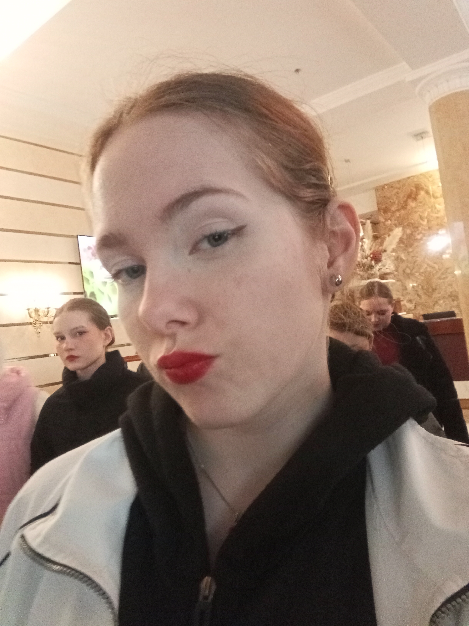 The image presents an indoor scene located in Saint Petersburg, Russia. The foreground is dominated by a young female with light skin and reddish-brown hair pulled back, looking directly into the camera. She wears prominent white and black winged eyeliner, bright red lipstick with pursed lips, and a small stud earring in her left ear. Her attire consists of a black hooded sweatshirt layered under a white jacket with a visible metallic zipper detail; a thin necklace is also visible.
In the mid-ground and background, several other young females are present. To the left, a female with dark hair pulled back, wearing red lipstick and eye makeup, is seen in a black top, looking away to her left. Towards the right background, another female with light-colored hair pulled back, dressed in a red top and black jacket, is partially visible, appearing to look downwards. A third female's head with lighter hair is partially obscured between the foreground subject and the background figures.
The environment features light-colored walls; a section on the left displays horizontal textured panels, while a section on the right is covered in a textured, possibly gold-toned, decorative material with visible floral or branch-like arrangements. A decorative wall sconce with multiple light sources is mounted on the left wall, and a screen displaying green and pink imagery is partially visible behind the leftmost background figure. The white ceiling exhibits decorative molding and is equipped with overhead lighting fixtures. A dark, possibly wooden, furniture piece or counter is present in the far right background. The overall setting suggests a waiting area or common room.