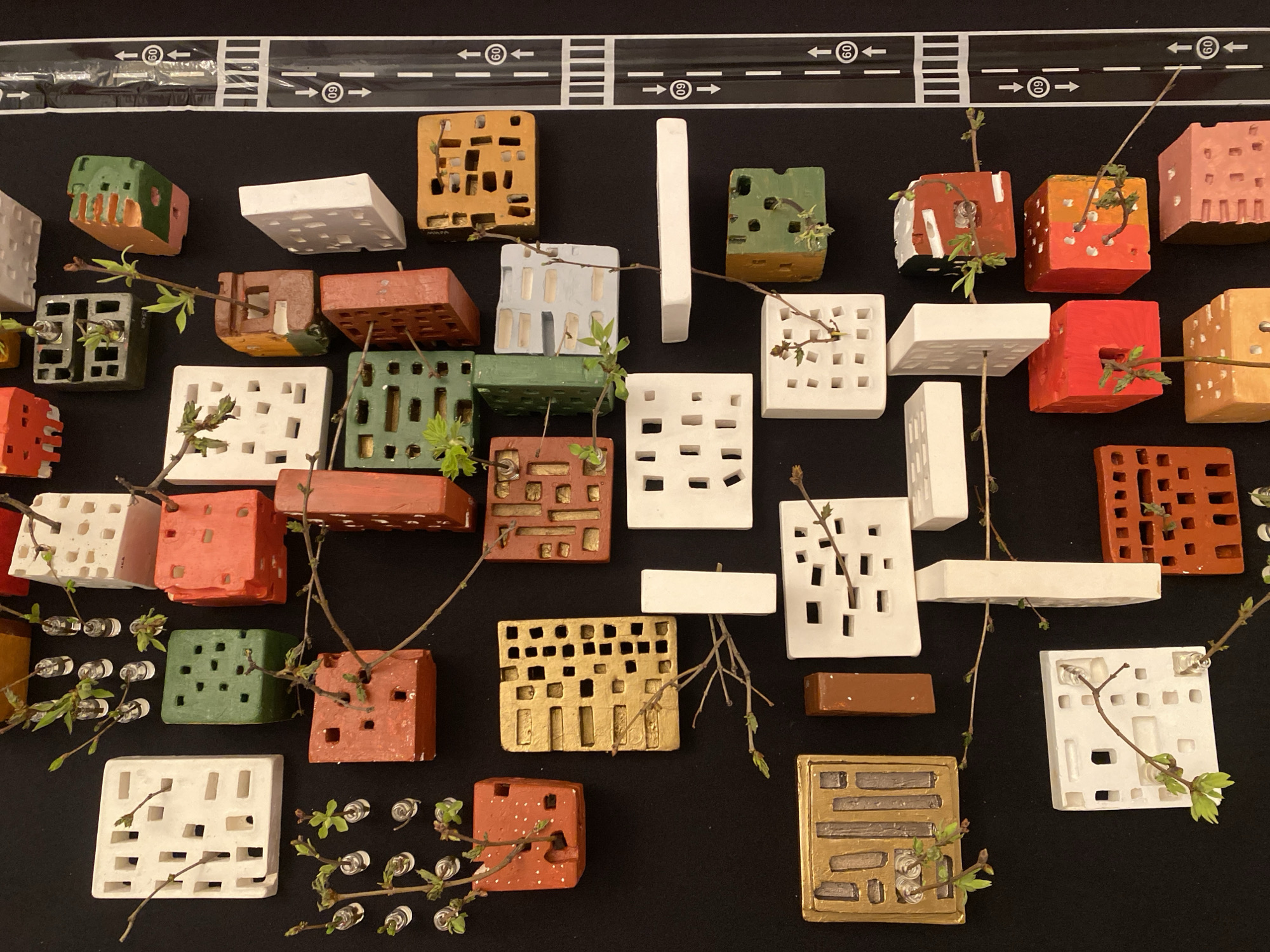 The image shows a collection of miniature building models arranged on a black surface, with a road sign design at the top edge of the frame. The models, varying in color and design, appear to be made of ceramic or similar material, featuring hollowed-out spaces and different geometric shapes. Many of the buildings have small, budding branches or twigs protruding from them, suggesting a theme of urban growth or nature reclaiming architecture. The presence of tiny glass vials with sprouts beside some of the buildings hints at an intentional arrangement. The scene is set against a plain, non-reflective backdrop, emphasizing the models. The road sign at the top indicates a road with speed limits, with white arrows indicating traffic flow direction. The location is not specified or evident from the image details.