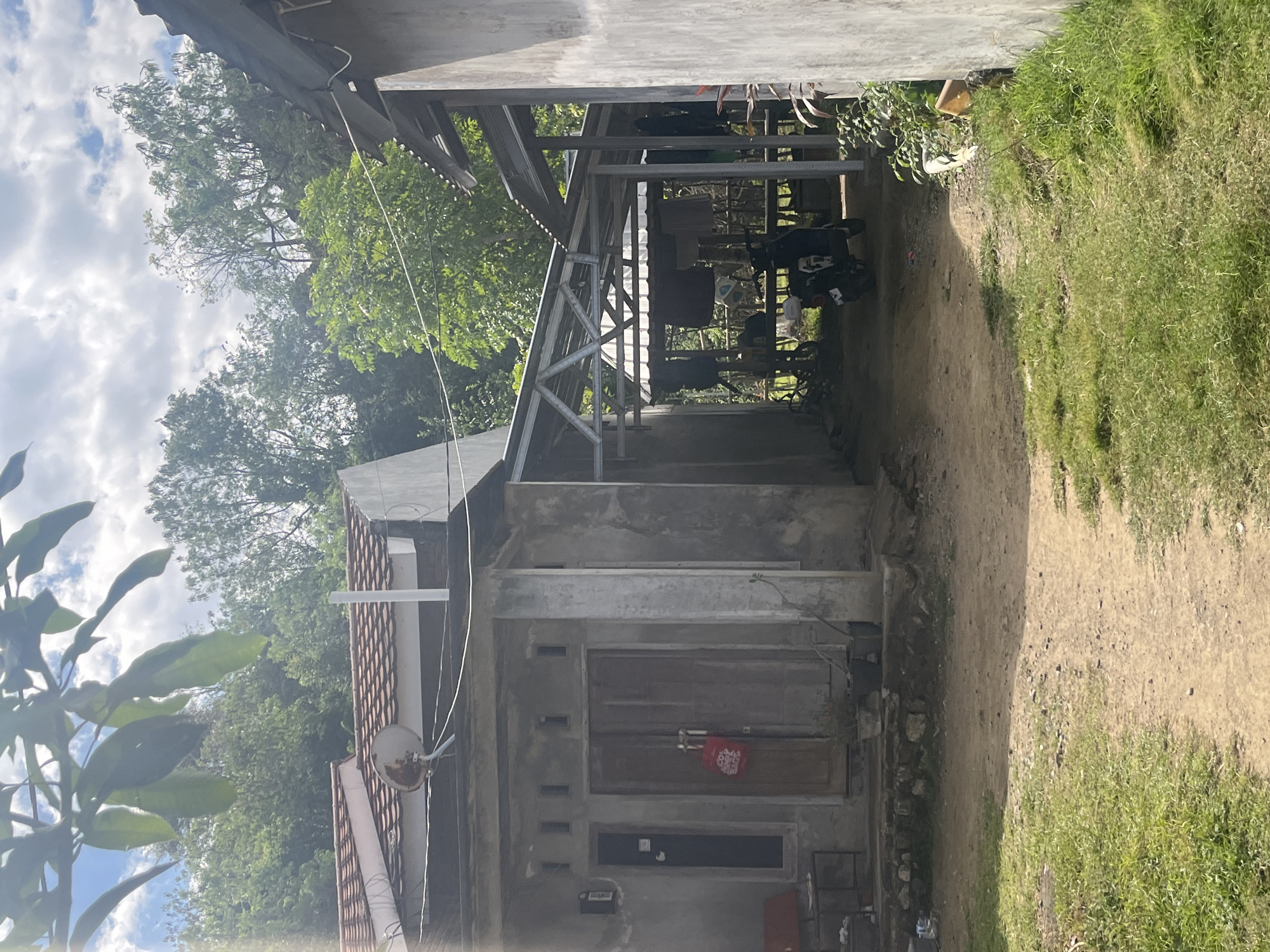 The image depicts a scene outside a simple residential structure, likely in a rural or semi-rural setting. In the foreground, a dirt path winds through patches of green grass. To the left, a small house with a tiled roof and concrete walls is visible. The house has a double wooden door with a red bag hanging on it, and small rectangular windows above the door. To the right, a neighboring structure with concrete walls and a corrugated metal roof extends forward, creating an open-air covered area. This area is used for utility purposes, with clothes hanging on a line and a motorcycle parked underneath. Lush green trees and foliage surround the structures, suggesting a natural or semi-natural environment. The sky above is partly cloudy. The overall impression is one of a humble dwelling in a tropical or subtropical climate.