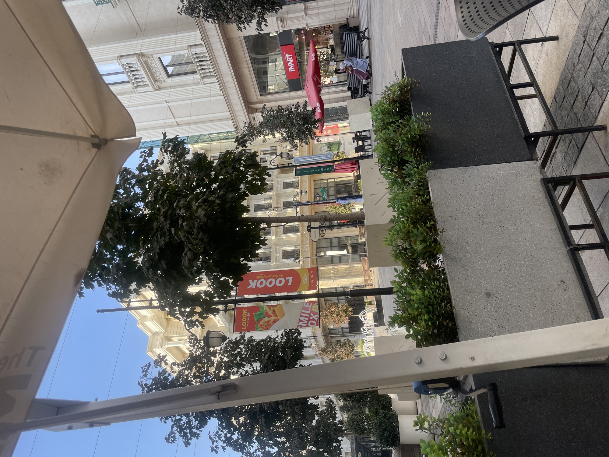 The image shows a wide pedestrian walkway in Tashkent, Uzbekistan, in front of an outdoor cafe and shops. The walkway is paved with grey stone tiles and features planters with green shrubbery. In the foreground, a portion of a large white umbrella and structural supports for an outdoor seating area are visible.
Further down the walkway, there are trees with dark green foliage and lampposts. Two large banners hang from one of the lampposts. The banner on the left displays the word "LOOOK" in red letters above an image of fried chicken. The banner on the right also says "LOOOK" in red letters with the slogan "We love your smile." Below these, another banner advertises "MILE OX" and provides a phone number.
Beyond the banners, there is a building with a light-colored facade, multiple windows, and decorative balconies. Further to the right, a store with a prominent red "iVART" sign is visible. An open red umbrella shades a bench where two women, dressed in headscarves and long clothing, are seated. A fountain is visible in the background, with jets of water. The sky is clear and blue. The overall scene suggests a pleasant day in a commercial or public area.