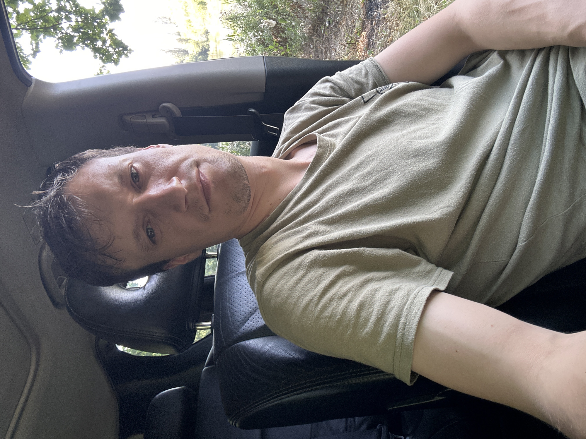 A man sits in the driver's seat of a vehicle, facing the camera. He is wearing a light green t-shirt. The interior of the car, including a black leather seat and headrest, is visible. Outside the car, through the open window and windshield, there is a view of trees, foliage, and a body of water, suggesting a rural or natural setting. The lighting indicates it is daytime.