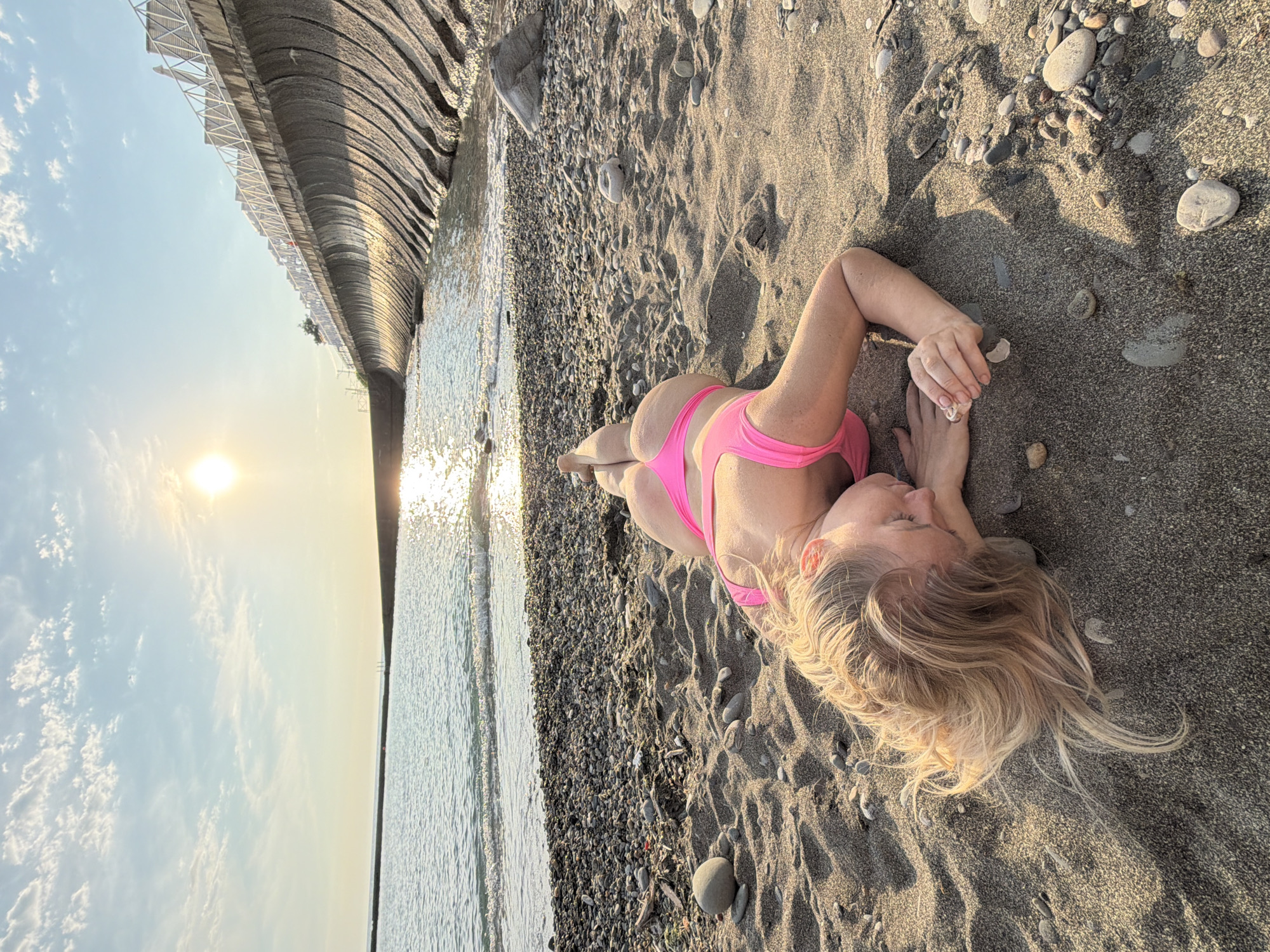 A blonde woman in a pink bikini lies on her stomach on a sandy beach in Sochi, Russia, with her head resting on her arms. The sun is setting over the Black Sea, casting a warm glow on the scene. In the background, a wave-breaking wall with a white railing lines the shore. The beach is covered in dark sand and pebbles.