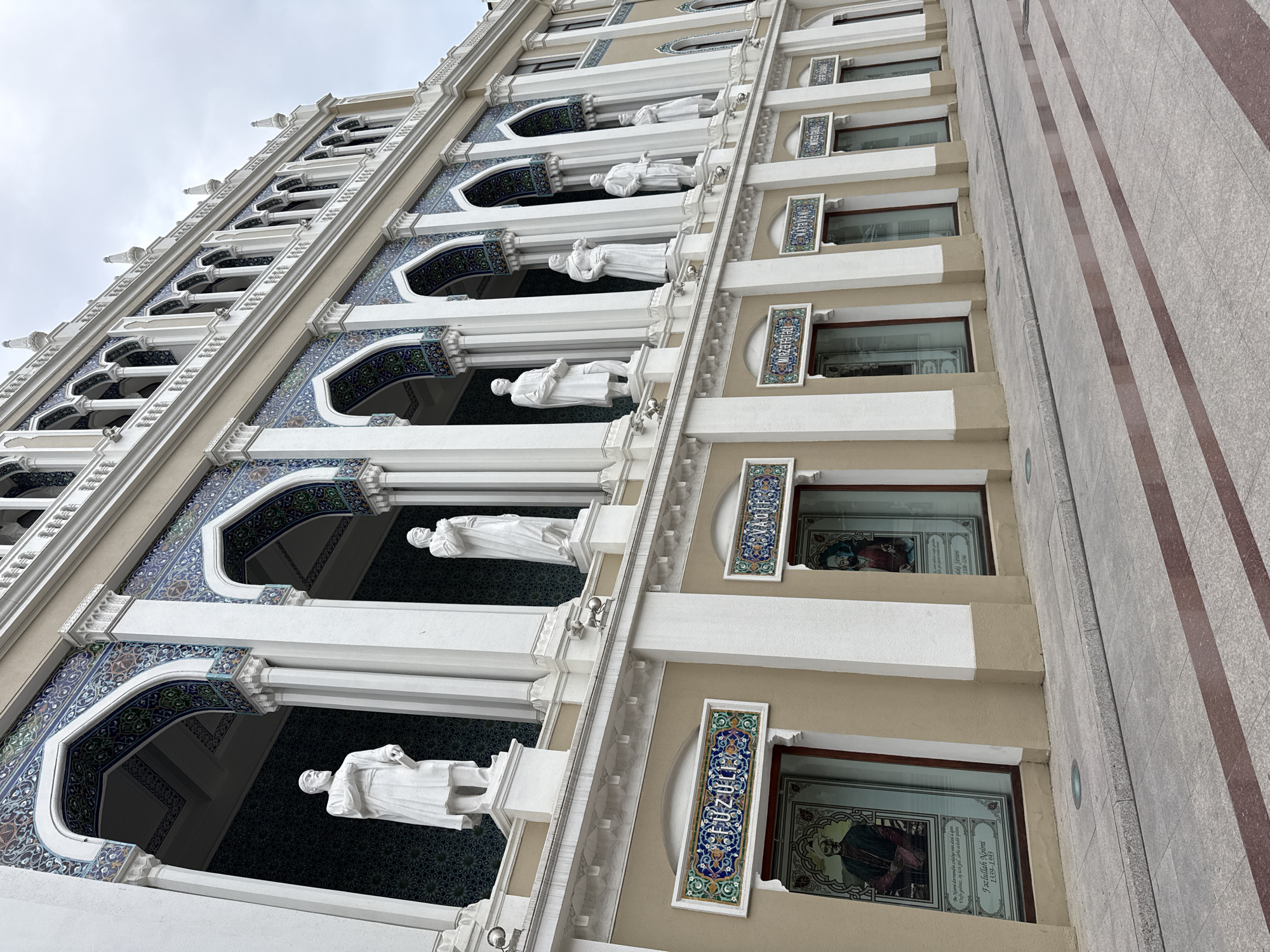 This image showcases the exterior of a building, likely a mosque or a historical site, with a focus on its architectural details and decorative elements. The building features multiple levels with arched openings, adorned with intricate blue and green patterned tiles. White statues of people in draped clothing are positioned within these openings, adding a classical or religious element. The building's facade is primarily a light beige color, contrasted by the ornate tile work and white architectural elements.
Windows on some levels are visible, some containing what appear to be stained-glass images or portraits. The OCR text visible on these windows refers to individuals named "Fazlullah Naimi" (1339-1393) and "alaki Siruni" (1108-1146), suggesting historical or religious significance. One window also contains Persian script, which translates to "We have sent you down from God's presence, as you have brought life to the world. The righteous are granted victory, by God's will."
The perspective is from a low angle, looking up at the building, which emphasizes its height and grandeur. The sky is overcast, indicating a cloudy day. To the right of the building, a textured stone pavement with reddish-brown stripes is visible, suggesting a walkway or plaza. The overall impression is one of ornate, possibly ancient, architecture with a clear cultural or religious context, set against a muted sky. The location is identified as Sabail Raion, Azerbaijan.