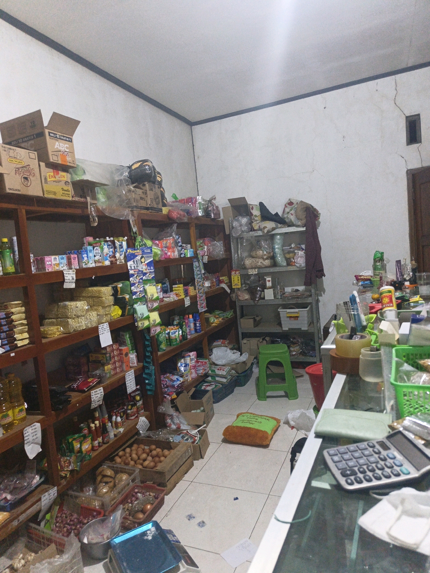 The image depicts the interior of a small convenience store, possibly a warung, located in Kamulyan, Indonesia. The store is filled with shelves stocked with various goods, including packaged food items, beverages, cleaning supplies, and some fresh produce like onions and eggs. Cardboard boxes are stacked on top of some shelves. A green plastic stool is placed near a metal shelving unit. On the floor, there is an orange pillow shaped like toast and some plastic bags. In the foreground, a counter is visible with a calculator and other assorted items. The walls are white and show signs of wear, with visible cracks. The lighting appears to be artificial, casting shadows across the space. There are no people visible in the image.