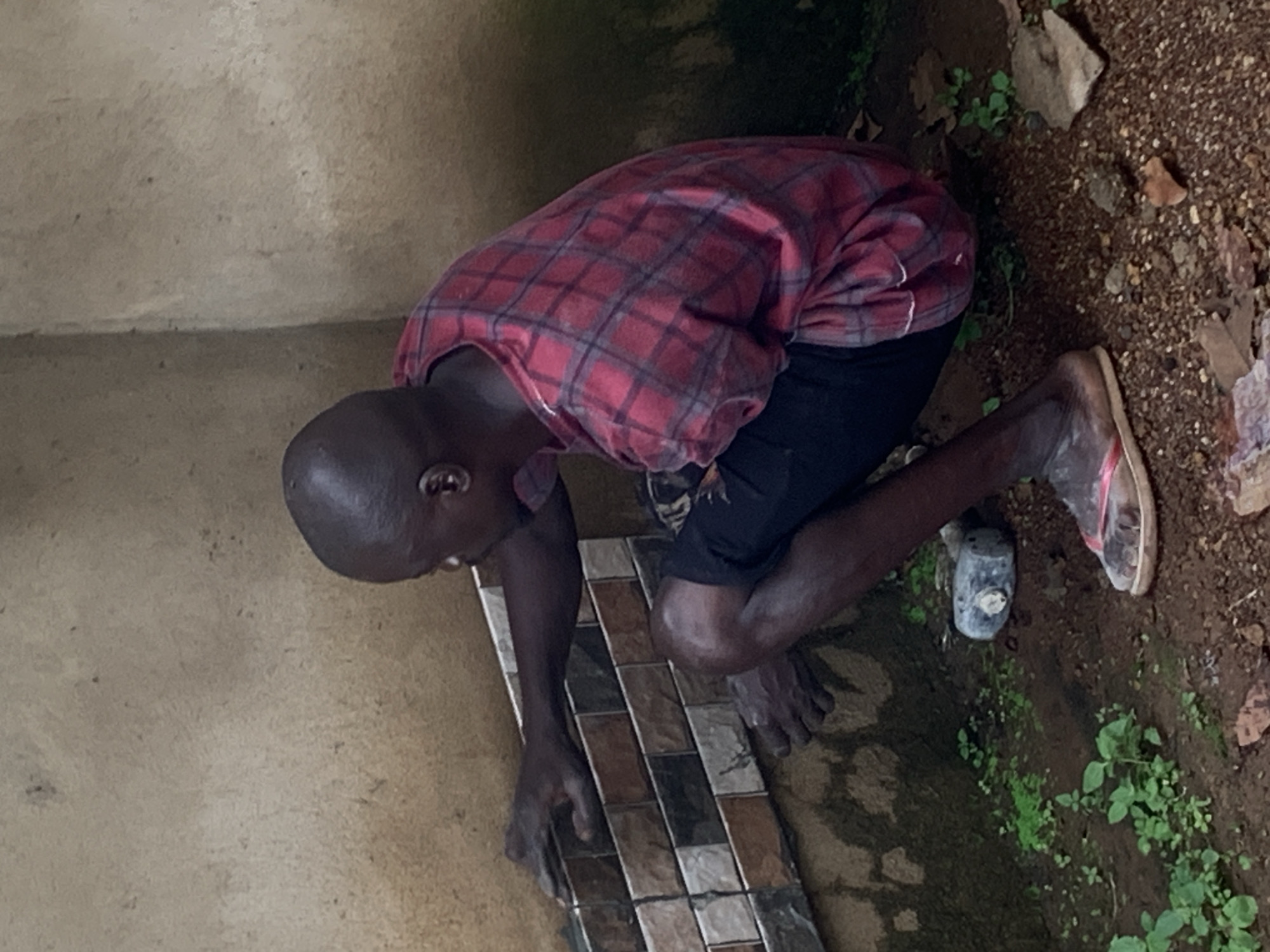A man with dark skin, bald head, and dark clothing is crouching and tiling a wall with rectangular tiles. He is wearing a red and black plaid shirt and black shorts. His feet are bare except for a pair of worn flip-flops. The wall is made of rough, light-colored plaster, and the floor is dirt with small green plants growing. A grey object, possibly a container, is on the ground next to the man's feet. The scene appears to be outdoors, possibly in a rural or developing area. The location is identified as Agban, Nigeria.