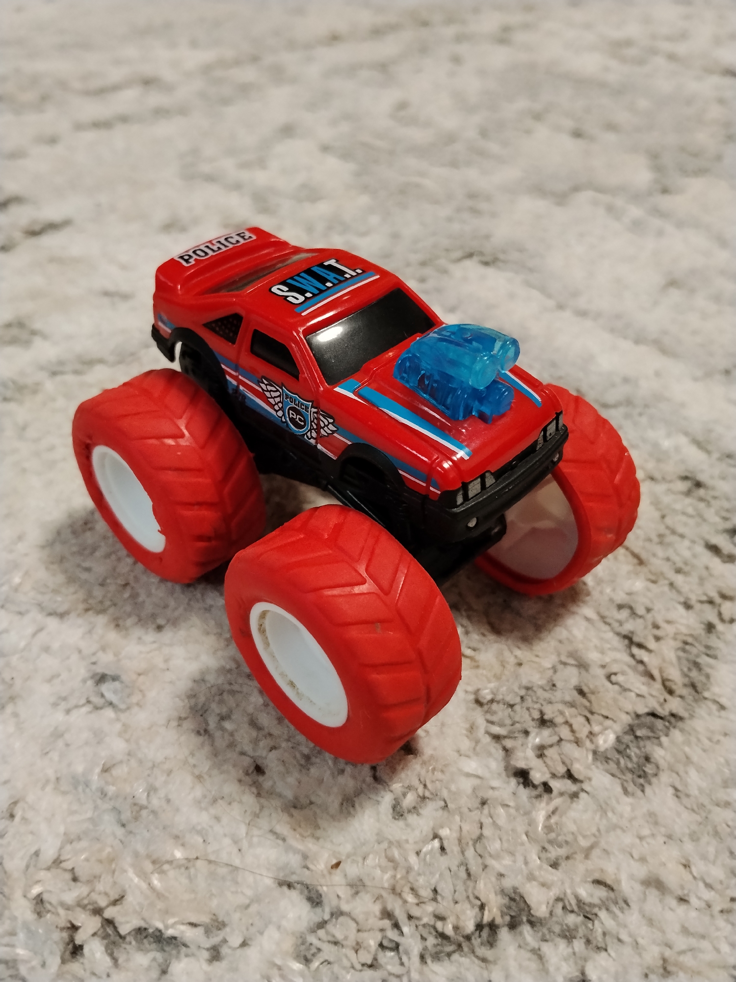 A red toy monster truck with "POLICE" and "S.W.A.T." markings is positioned on a textured, light-colored surface, likely carpet or a similar fabric. The truck features large, red tires with white rims, a blue engine intake on the hood, and blue and white racing stripes along its body. The background is blurred, indicating a shallow depth of field. There are no people or identifiable environmental features of a city present in the image. The image does not provide any information about the city of Буренка, Russia.