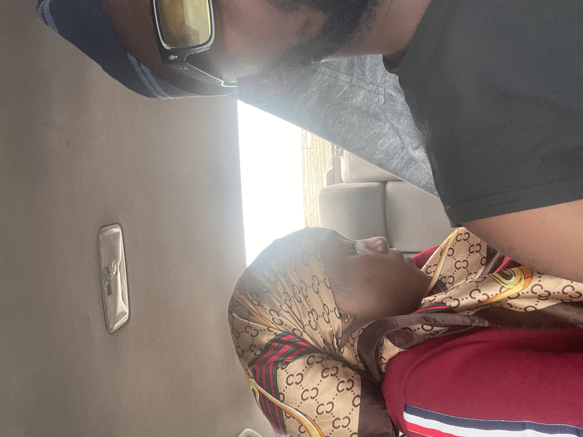 The image shows two people, a man and a woman, in what appears to be the interior of a vehicle. The man is on the left side of the frame, wearing sunglasses and a blue cap. He has a beard and is wearing a dark t-shirt. The woman is on the right, wearing a headscarf with a Gucci-like pattern and a maroon top. She is looking away from the camera, with her head tilted back. The interior of the vehicle is dimly lit, with a light fixture visible on the ceiling. The background suggests an outdoor scene with some furniture, possibly in a residential area in Kwali, Nigeria. The overall mood is relaxed and casual, with the focus on the two individuals. The time of day is not explicitly indicated, but the lighting suggests it could be daytime.