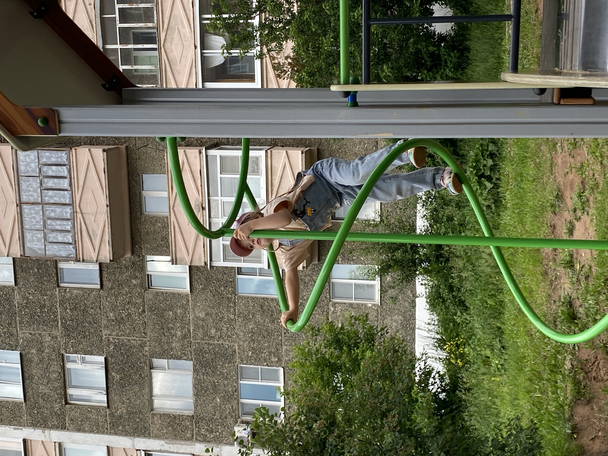 A young child, wearing a light brown t-shirt, blue denim overalls, and a pink baseball cap, is climbing on a green spiral slide structure in an urban playground. The child's feet are positioned on the lower part of the slide, and their hands are gripping the railing. Behind the playground equipment, a multi-story, brutalist-style apartment building with numerous windows and balconies is visible. The building's facade is made of rough concrete. Lush green trees and vegetation surround the playground, suggesting a warm weather environment. The overall scene depicts a typical day in a residential area of Votkinsk, Russia, where children play outdoors near their homes.
