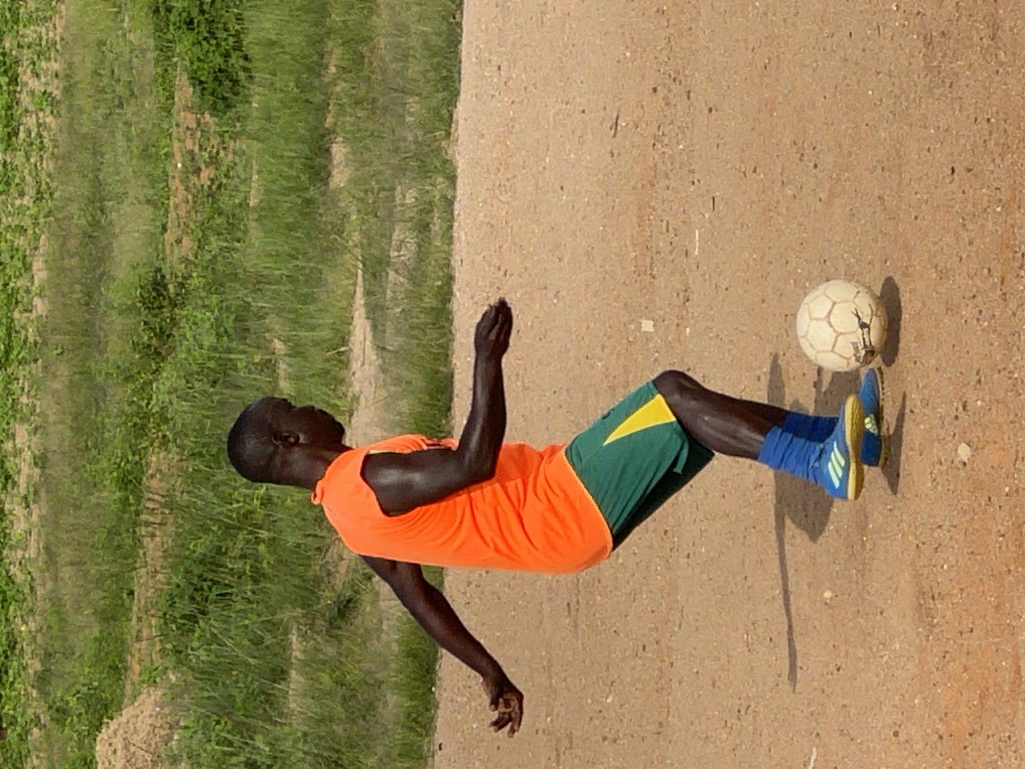 In Jos, Nigeria, a dark-skinned male is pictured playing soccer on a dirt field. He is wearing an orange t-shirt and green shorts with a yellow stripe. His blue socks and cleats are visible. The soccer ball is to his right. The background consists of green vegetation and a sandy area.