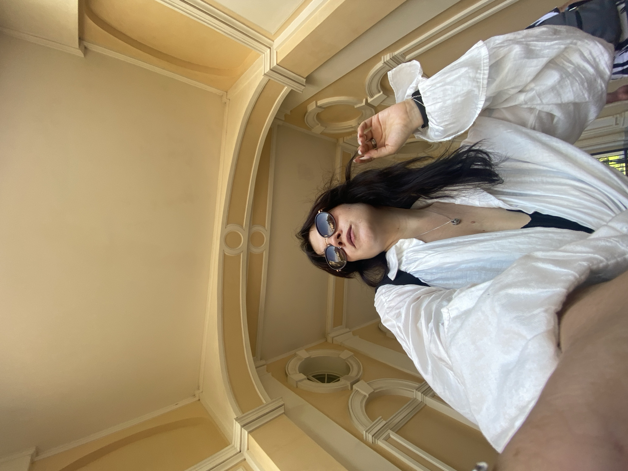 A low-angle, eye-level shot captures a young woman standing outdoors in an arched passageway with cream-colored walls and ceiling. She has dark hair, sunglasses with reflective lenses, and is wearing a white, billowy blouse over a black garment. Her left hand is raised, touching her hair. The architecture features ornate, rounded arches and decorative circular insets. The lighting suggests it is daytime. This location is identifiable as the Oranienbaum Palace complex in Lomonosov, Russia, specifically within one of its garden pavilions or arcades.