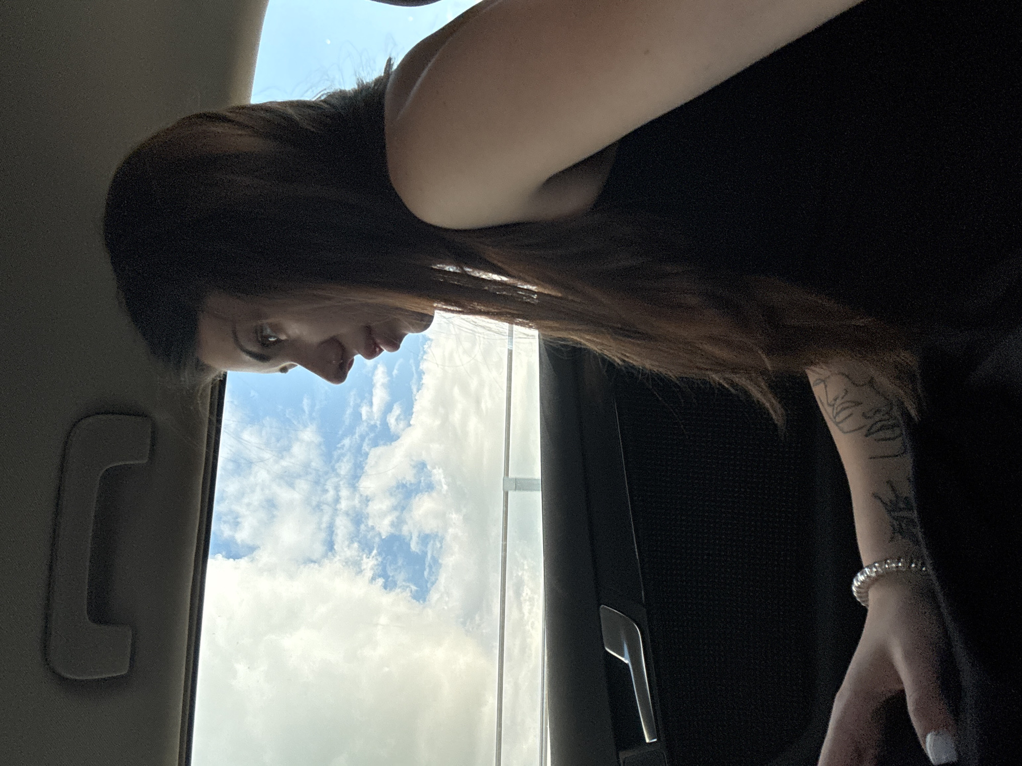 The image depicts a person inside a vehicle, captured from a slightly elevated angle looking down. The subject, seemingly a female, is seen in profile with long, dark hair, looking out a window. She is wearing a black top. Her left forearm is visible, adorned with a line art tattoo, and a silver-toned, possibly beaded, bracelet is worn on her wrist. Her fingernails have white polish.
The interior of the vehicle is partly visible, showing a light-colored headliner and a grey grab handle to the left of the person. A dark car door panel and window frame are also visible on the right. Through the window, a bright blue sky with scattered white, fluffy clouds is seen, indicating daytime with good weather. The scene suggests the person is traveling or situated in a parked car in Mramor, Bulgaria. There is no visible text in the image.