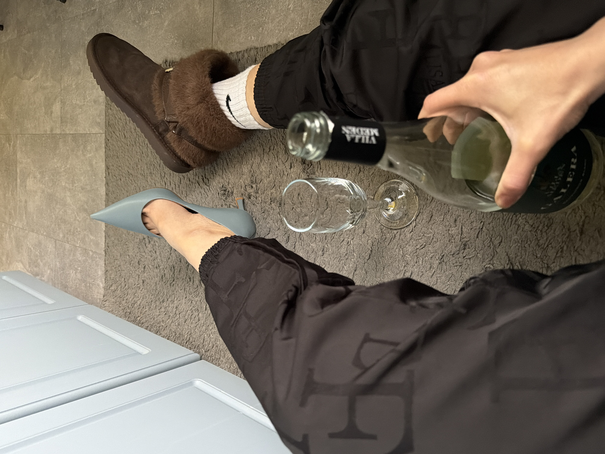The image presents an indoor scene. A person is visible, dressed in black pants, one leg adorned with a brown Ugg boot and white sock, while the other wears a light blue high-heeled shoe. A hand is captured pouring wine from a bottle labeled "VILLA MEDEN" into a wine glass. The setting includes a light-colored wall and a textured gray rug on the floor. No explicit evidence suggests this photo was taken in Pleven, Bulgaria, or any other specific location.