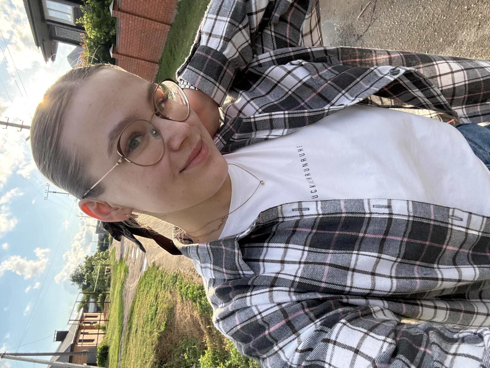 This is a vertical, close-up selfie of a young woman taken outdoors. The woman has blonde hair pulled back into two braids and is wearing glasses with thin, gold-colored frames. She is dressed in a white t-shirt with black text on the chest and a gray, black, and pink plaid open-front shirt over it. The text on her t-shirt appears to be in German, reading "KEHRUNRUHE". She is looking at the camera with a slight smile.
The background suggests a rural or suburban setting. There are houses with brick and dark siding visible, along with green grass and trees. A dirt or gravel path runs through the scene. The sky is blue with scattered white clouds, and the sun is low in the sky, casting a warm glow and creating a lens flare on the left side of the image. This indicates it is either early morning or late afternoon. The lighting suggests the weather is clear and pleasant. The woman's relaxed pose and smile convey a casual and happy mood.
