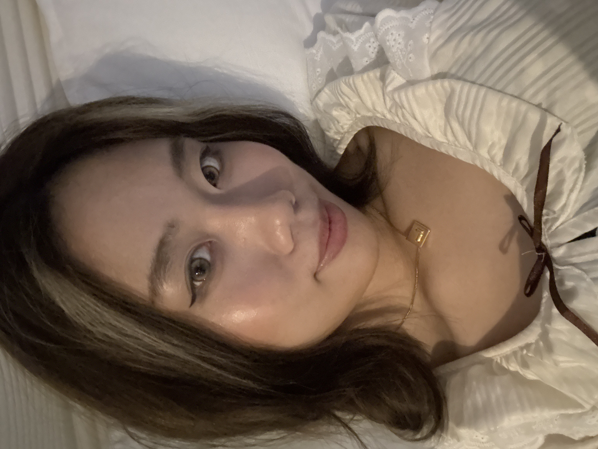 A woman is lying down on a white surface, likely a bed, wearing a white top with frills and a dark ribbon tie. She has brown hair with lighter streaks and is wearing eyeliner. A gold necklace with a small square pendant is visible. The background is blurred and out of focus. There is no indication of the location being in Chenghua District, China.