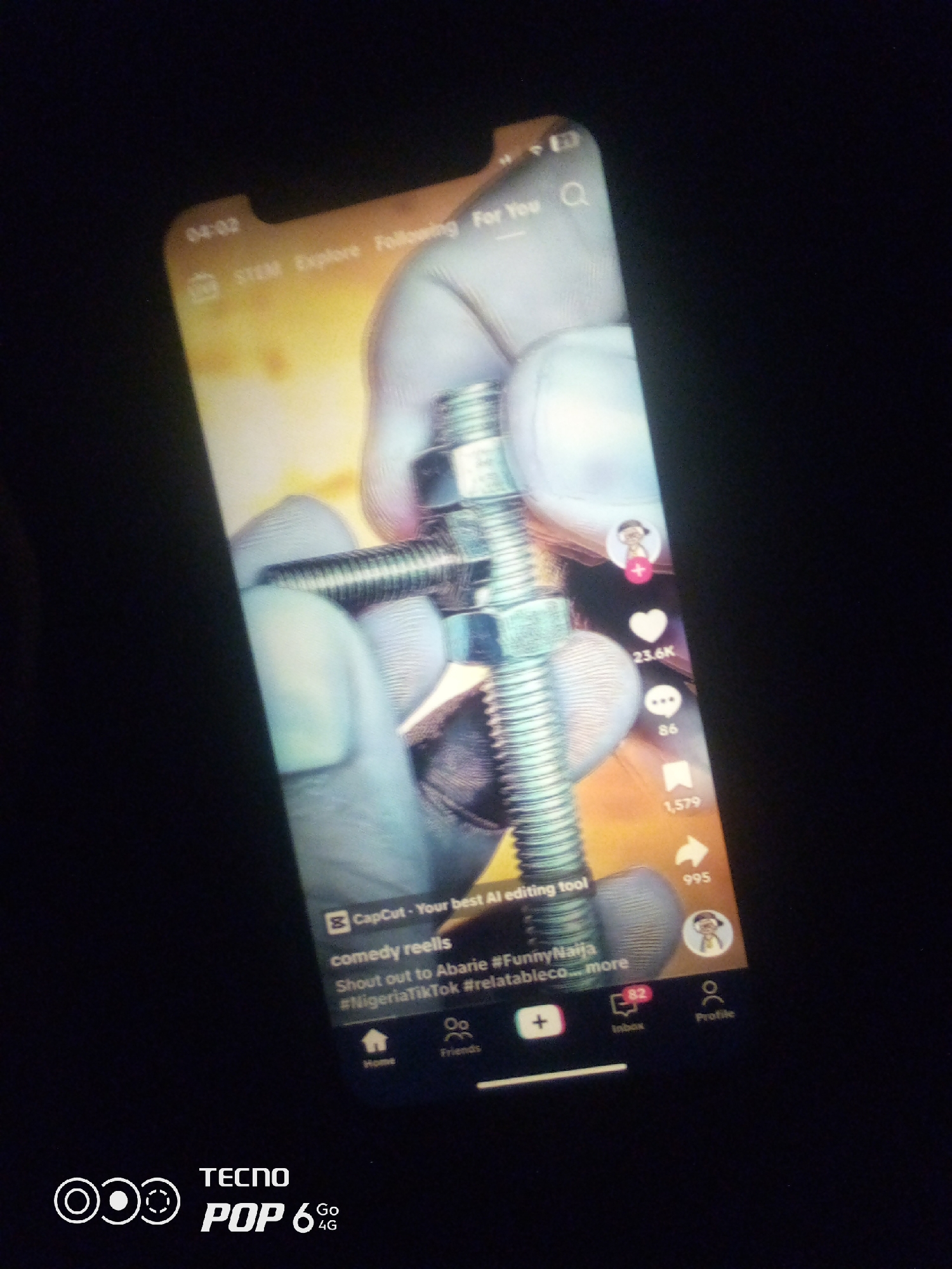 A phone screen displays a TikTok video of hands in light blue gloves assembling a bolt and nut. The video is from the "comedy reels" category, with hashtags like #NigeriaTikTok. The time on the screen is 04:02. On the right side of the screen, there are icons for likes (23.6K), comments (86), shares (1,579), and a user profile. At the bottom of the screen, a navigation bar shows icons for Home, Friends, a plus sign, Inbox (with a notification bubble of 82), and Profile. The bottom left corner of the image shows a TECNO POP 6 4G logo. The background of the image is dark, suggesting the photo was taken in low light conditions. There is no discernible information about the city of Howayi, Nigeria, within the image.
