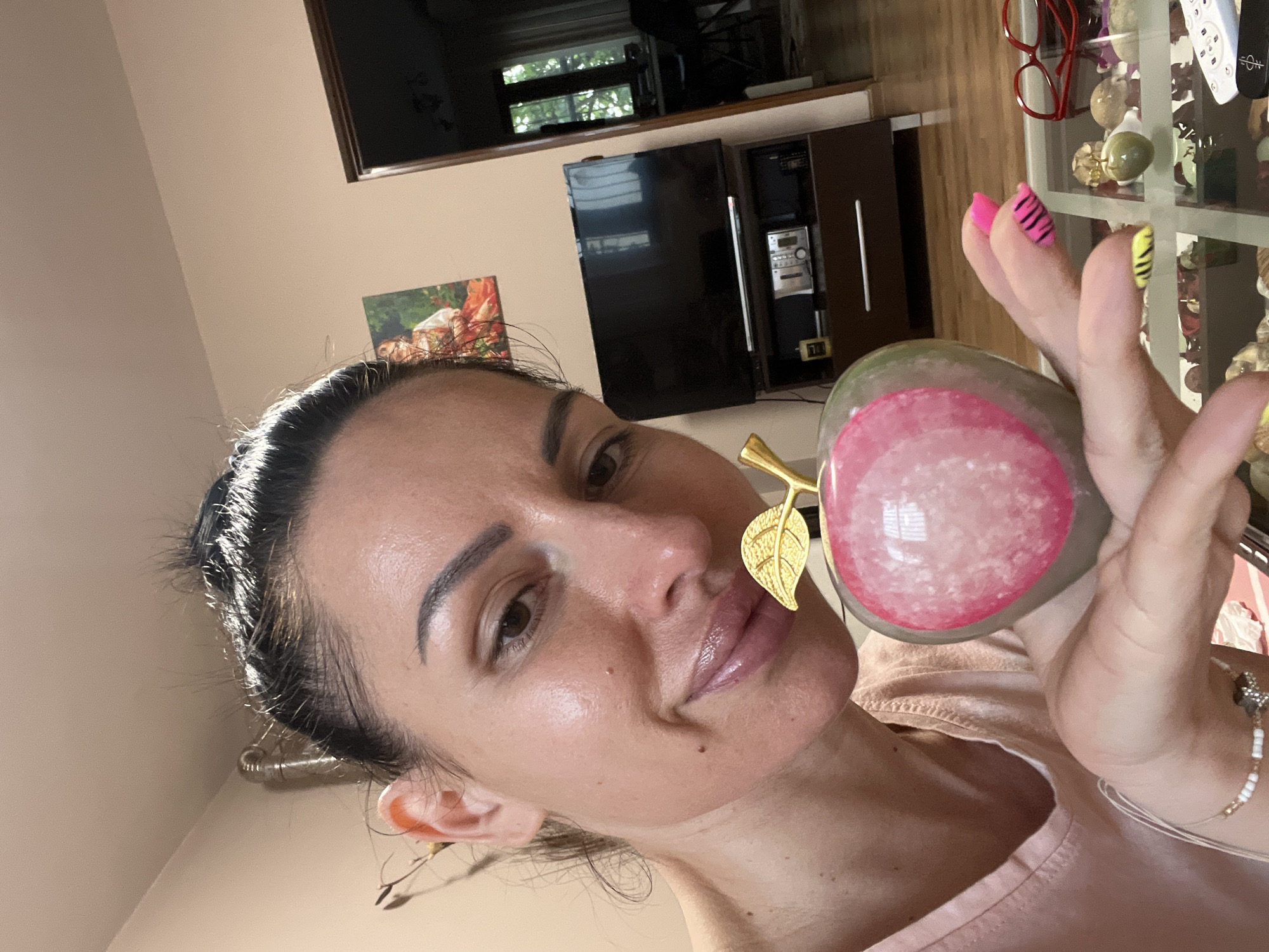 An image taken in Stara Zagora, Bulgaria, shows a woman holding up a decorative object, likely captured as a selfie. The woman, with dark hair pulled back and a natural makeup look, is smiling subtly as she looks towards the camera. She wears a light-colored, possibly pink, casual top.
In her right hand, she holds a polished, fruit-shaped object, possibly a stylized apple. It features a green or grey base with a prominent circular pink section, which has a textured, white mottled pattern within it. A small, shiny golden leaf is attached to the top of the object. Her nails are distinctively manicured; the thumb and index finger nails are neon pink with black zebra stripes, while her middle finger nail is yellow with black zebra stripes. A delicate beaded bracelet is visible on her wrist.
The background reveals an indoor setting, likely a living room, bathed in natural daylight. A light beige wall is visible behind her. To the left, a framed picture hangs on the wall. In the center, a dark brown entertainment unit holds a television and a stereo system. A window is visible above the television, showing greenery outside. To the right, behind the woman's hand, is a glass display cabinet containing numerous spherical and oval decorative objects, some appearing to be stone or glass, in various shades like green and beige. A pair of red-framed glasses also rests on this cabinet. No discernible text is visible in the image.