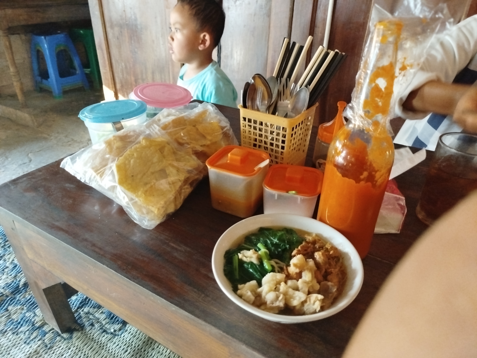A young boy with dark hair, wearing a light blue t-shirt, looks to the left, seated at a dark wooden table in a room with wooden walls and a wooden table. In front of the boy, on the table, is a white bowl of food containing noodles, green leafy vegetables, and pieces of meat. To the right of the bowl is a bottle of orange sauce, partially wrapped in plastic. Next to the bottle is a glass of dark liquid. A basket filled with chopsticks and cutlery sits behind the bowl. Several plastic containers with orange lids are also visible on the table, along with a bag of fried snacks. In the background, a blue plastic stool and a green plastic stool are partially visible. The floor is covered with a blue and white patterned mat. The scene appears to be a casual meal setting in Pojok, Indonesia.