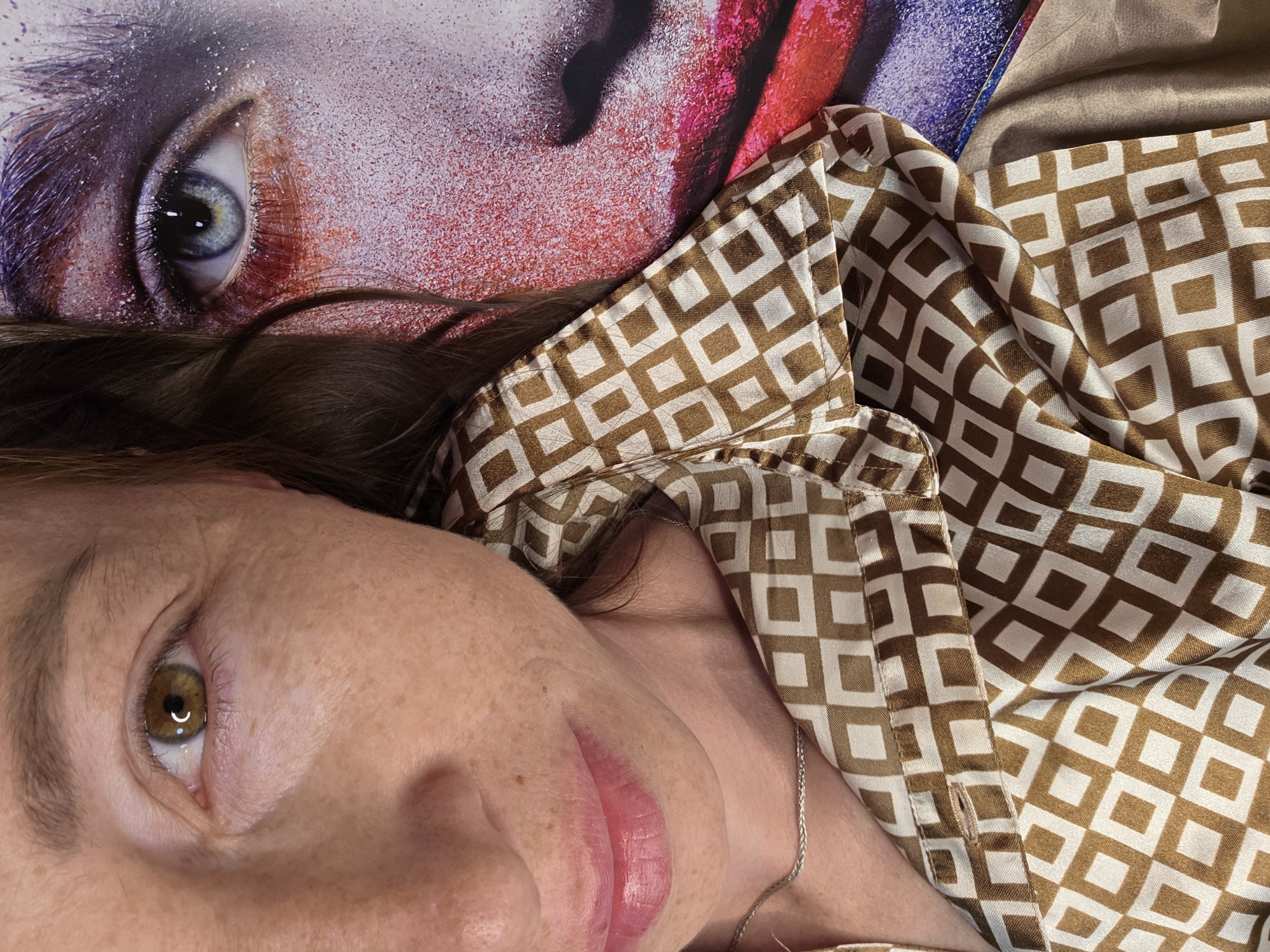 The image shows a close-up, overhead view of a woman's face and chest, positioned to the left of a large, abstract print of a face with dramatic eye makeup. The woman's face is partially visible, with her right eye and lips in focus. Her right eye is brown with a distinct highlight, and her skin has visible freckles and some fine lines. Her lips are slightly parted and appear to have a glossy pink lipstick. She is wearing a brown and white diamond-patterned shirt and a thin silver chain necklace. Her hair, which is brown, partially obscures the print on the right side of the image. The print itself features an eye with blue and green irises, surrounded by vibrant red and purple makeup and glitter. The overall composition is intimate and focused on the textures and colors of both the woman and the artwork. The location is unknown.