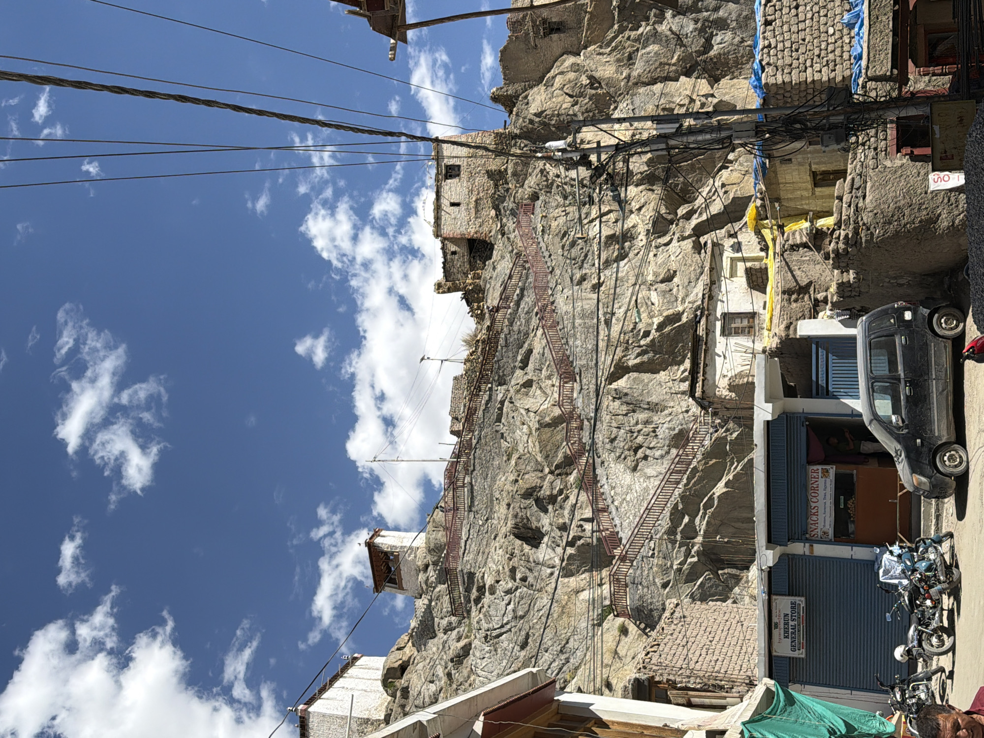 The image captures a street scene in Leh, India, with a steep, rocky mountainside dominating the background. The mountain is dotted with several old, stone structures, some of which appear to be monasteries or fortresses. A prominent feature is a long, red-railed staircase ascending the mountainside, leading to a white building with a square tower at the summit. The sky above is a vibrant blue, with scattered white clouds.
In the foreground, a row of shops lines the street. One shop, labeled "SNACKS CORNER," advertises momos, samosas, tea, and thukpa. Another shop is identified as "KHERUN GENERAL STORE." Several motorcycles are parked in front of the shops, and a black car is visible in the middle of the street. A man, partially visible in the bottom left of the frame, is wearing a red shirt. The scene is characterized by a mix of traditional architecture, a rugged natural landscape, and the everyday life of a bustling street. Utility poles with numerous electrical wires are also present, indicating the presence of modern infrastructure. The overall impression is one of a historic and vibrant town nestled within a dramatic natural setting.
