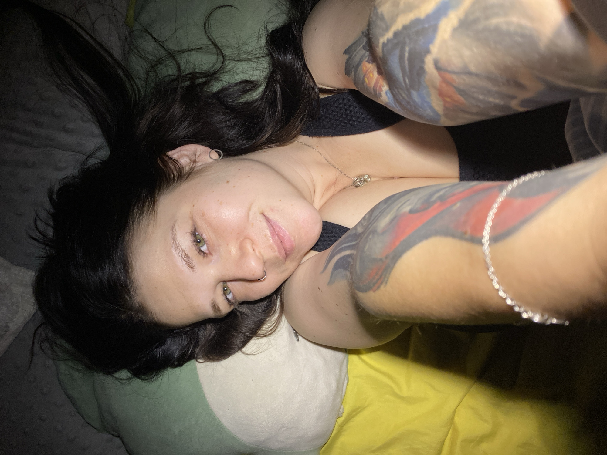 A woman with dark hair and tattoos on her arms is lying on a bed or couch. Her head rests on a green and white pillow. She is wearing a black top and a silver necklace. The environment appears to be indoors, possibly a bedroom. There is no verifiable information present in the image to indicate it was taken in Chaikovsky, Russia, or any other specific location.
