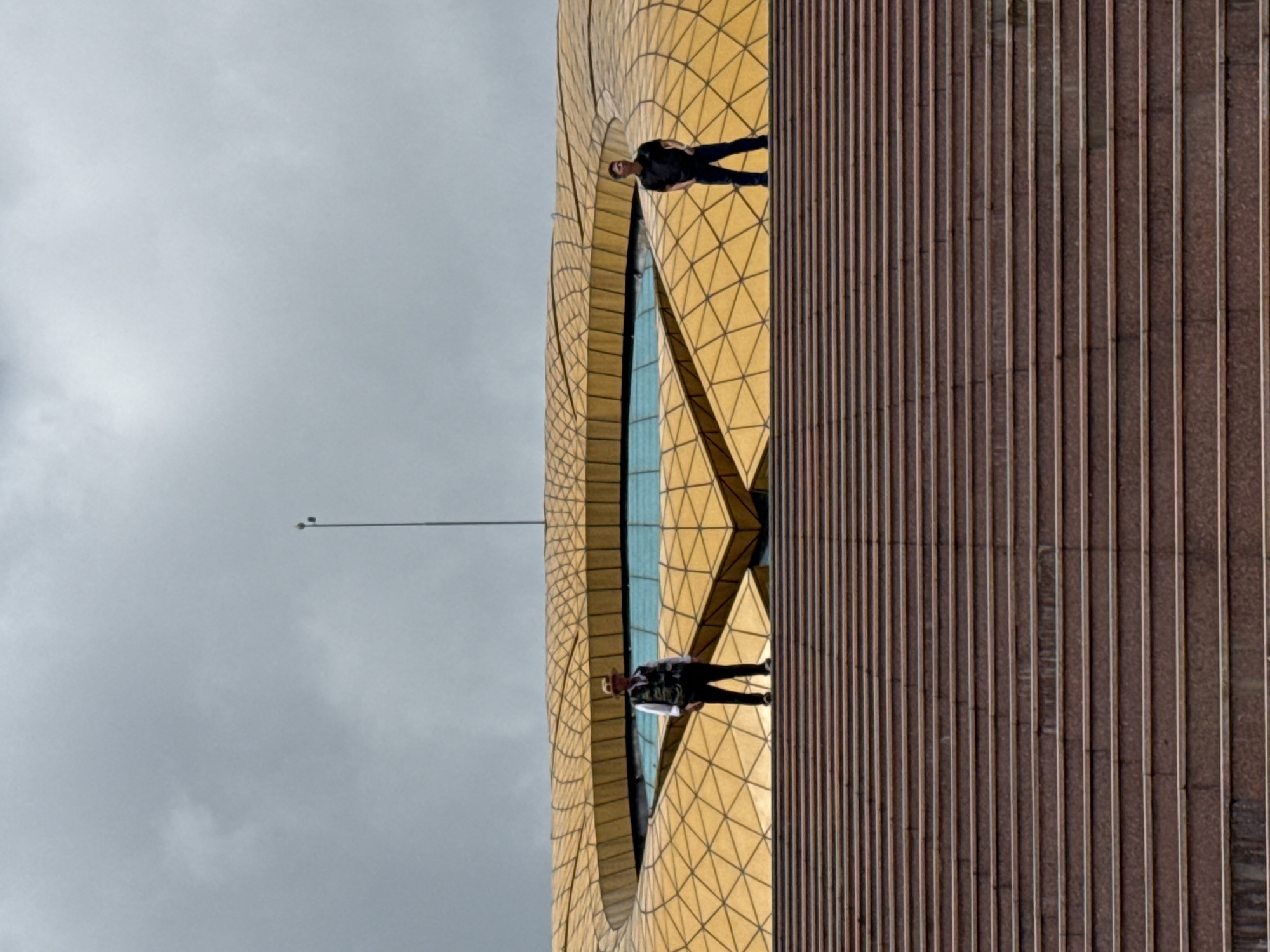 This image captures a wide, ascending staircase of reddish-brown stone leading to a distinctive modern building in Lam Vien Square, Da Lat, Vietnam. The building features a prominent golden roof, composed of numerous geometric facets, which surrounds a central, irregular-shaped blue glass skylight. The sky above is uniformly overcast and grey.
Two adult males are positioned on the uppermost visible step of the staircase. The man on the left wears a light-colored long-sleeved shirt, a dark vest, dark trousers, and a light-colored hat, facing directly forward. The man on the right wears a dark short-sleeved polo shirt and dark trousers, with his hands in his pockets, and is looking slightly to his right. Both individuals are standing still. A thin, dark pole, equipped with two small spherical attachments near its top, extends vertically from behind the golden building into the grey sky. The photograph is taken from a low angle, emphasizing the scale of the steps and the building.